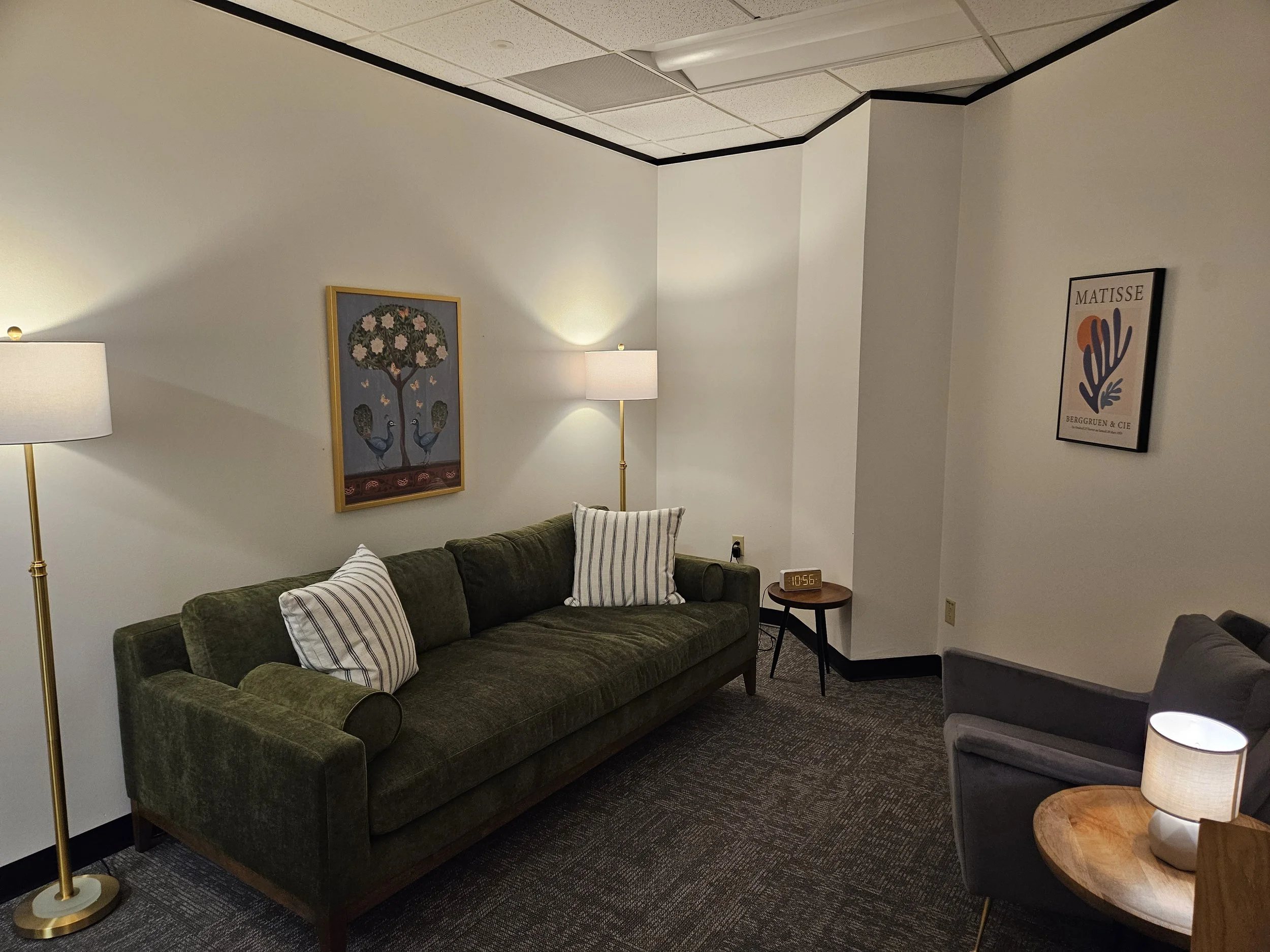 Living room with green velvet sofa, striped pillows, floor lamp, wall art of a peacock and tree with flowers, small round side table, an alarm clock, and a gray armchair.