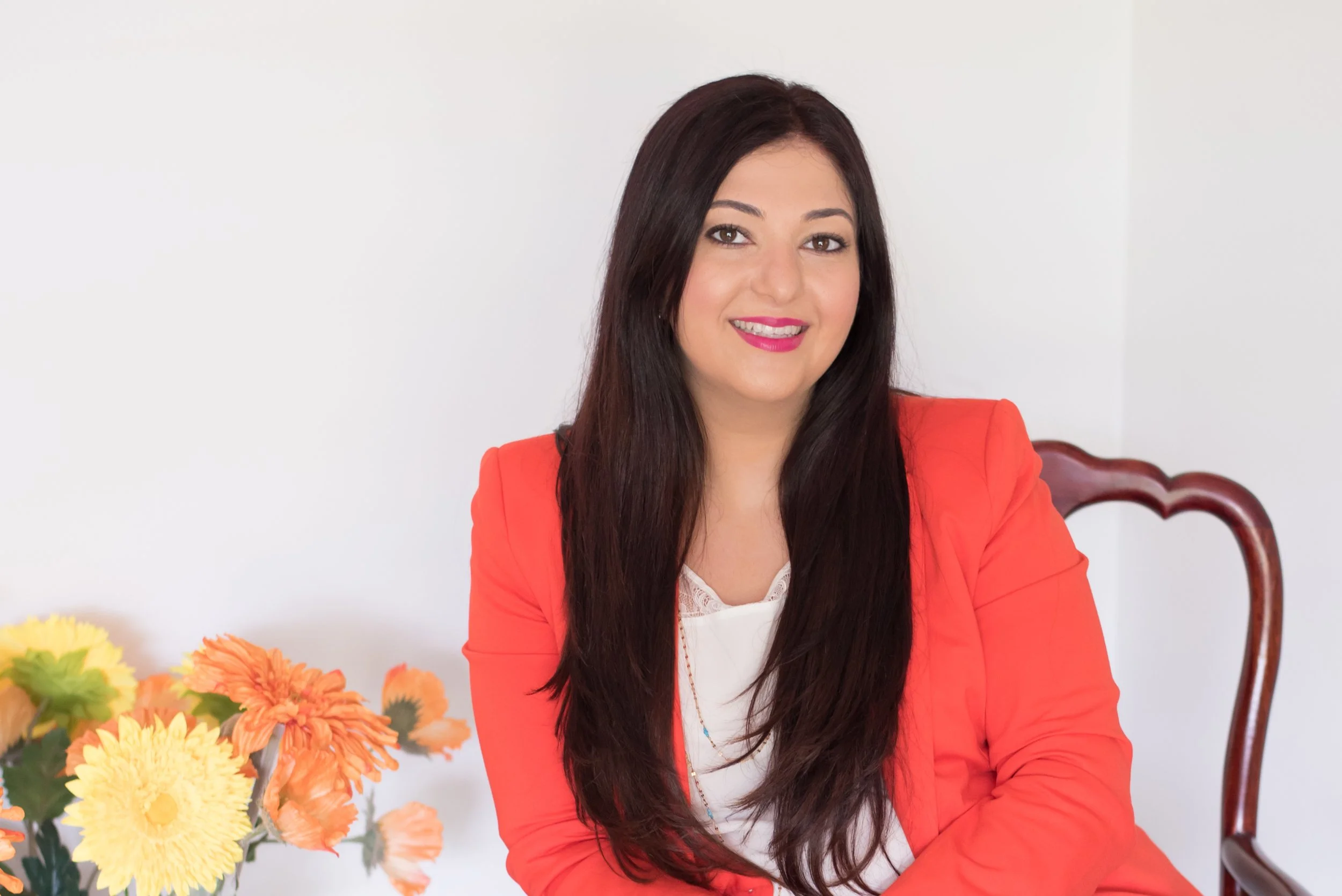 A woman with long dark hair, wearing a red blazer and white top, sitting at a table with orange and yellow flowers, smiling at the camera against a white background.