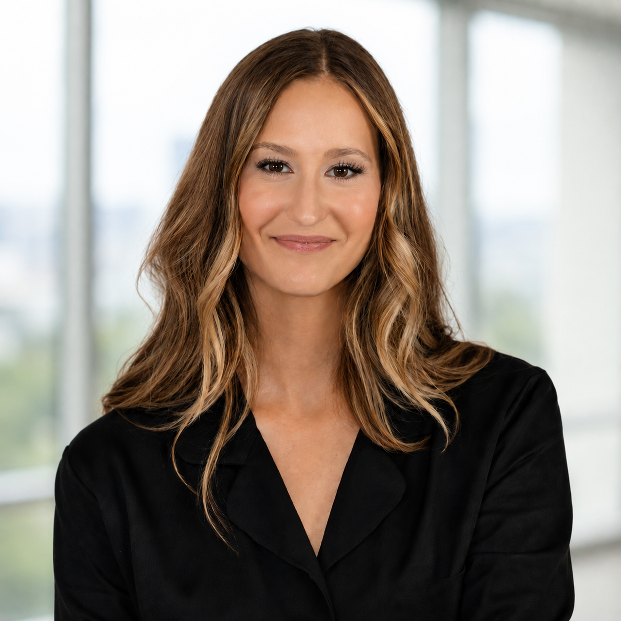 A woman with shoulder-length wavy brown hair wearing a black blazer, smiling in front of a blurred window background.