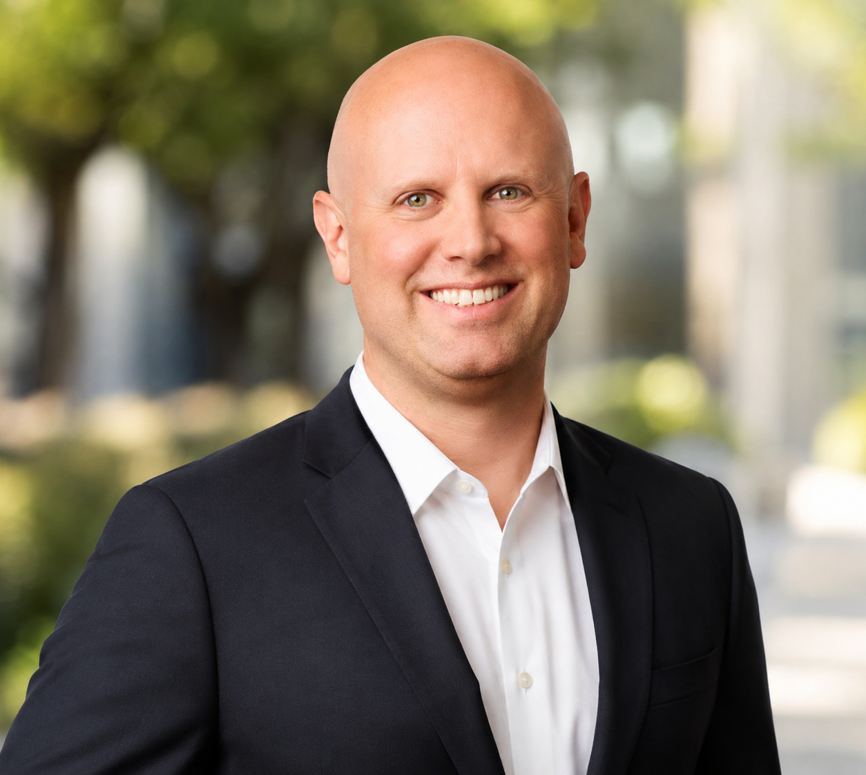 A smiling bald man wearing a black suit and white shirt, standing outdoors with blurred trees and buildings in the background.
