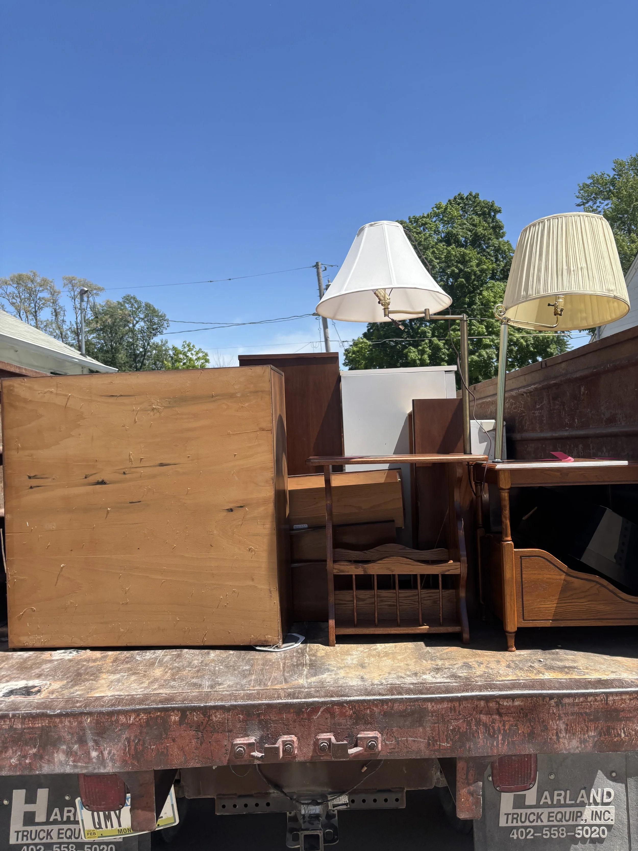 Various pieces of furniture including lamps, chairs, and shelves placed on a flat surface outdoors, with trees, power lines, and a blue sky in the background.