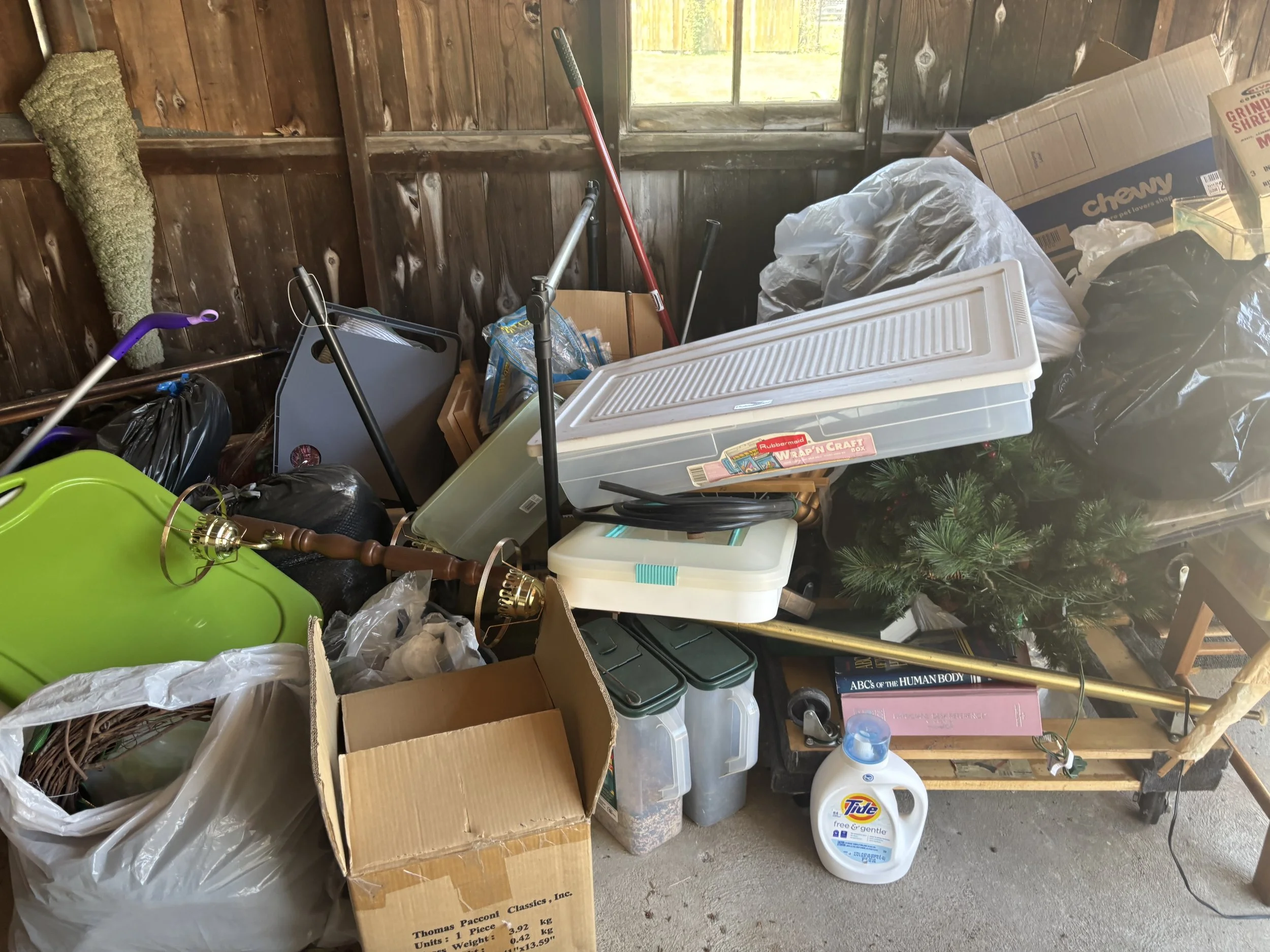 A cluttered garage with various items including cardboard boxes, plastic storage bins, a Christmas tree, cleaning supplies, a lamp, books, and tools.