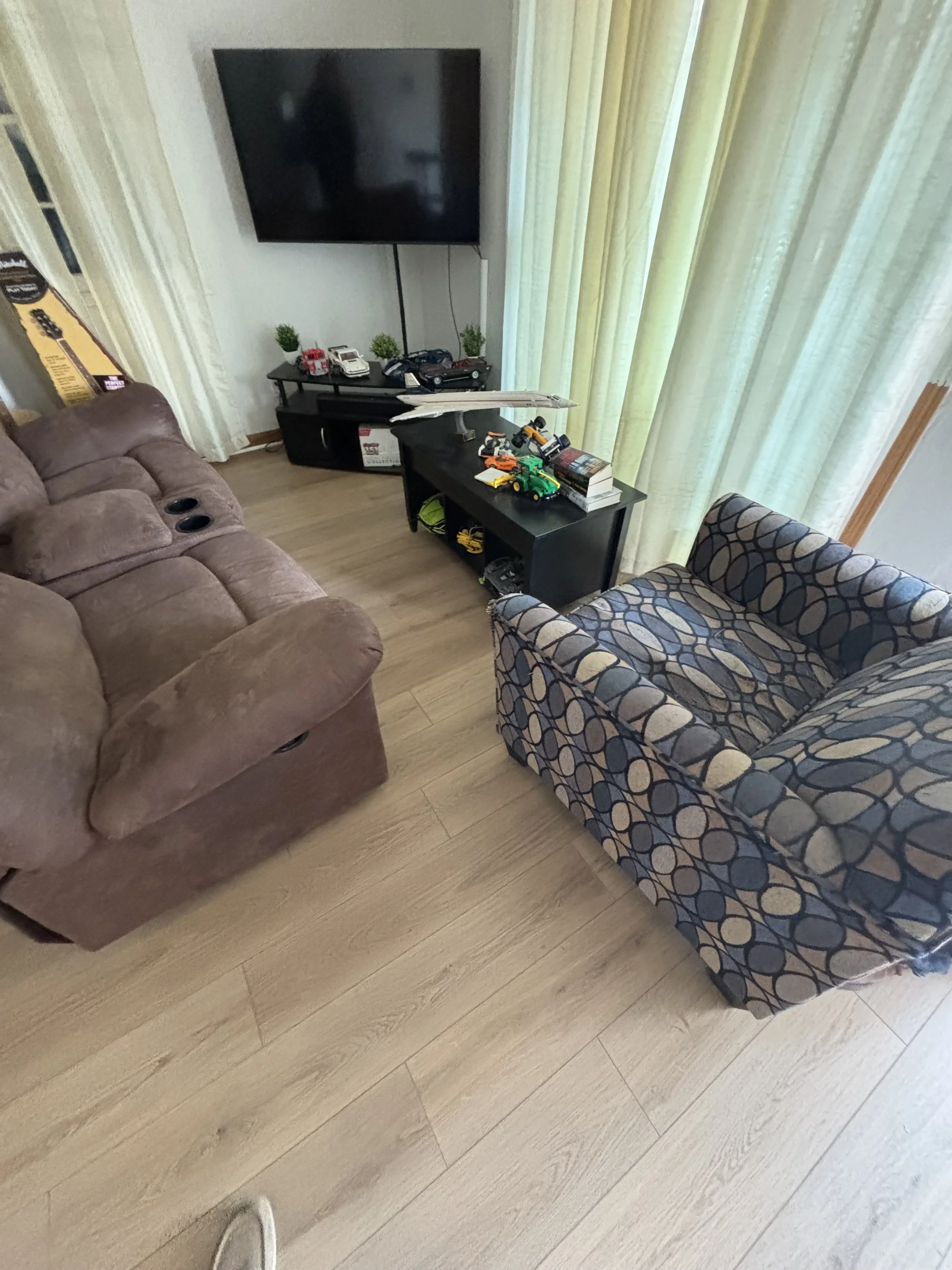 Living room with a brown plush sofa, a patterned blue and beige armchair, a black TV stand with toy cars, books, and decorative plants, and a wall-mounted flat-screen television with green curtains in the background.