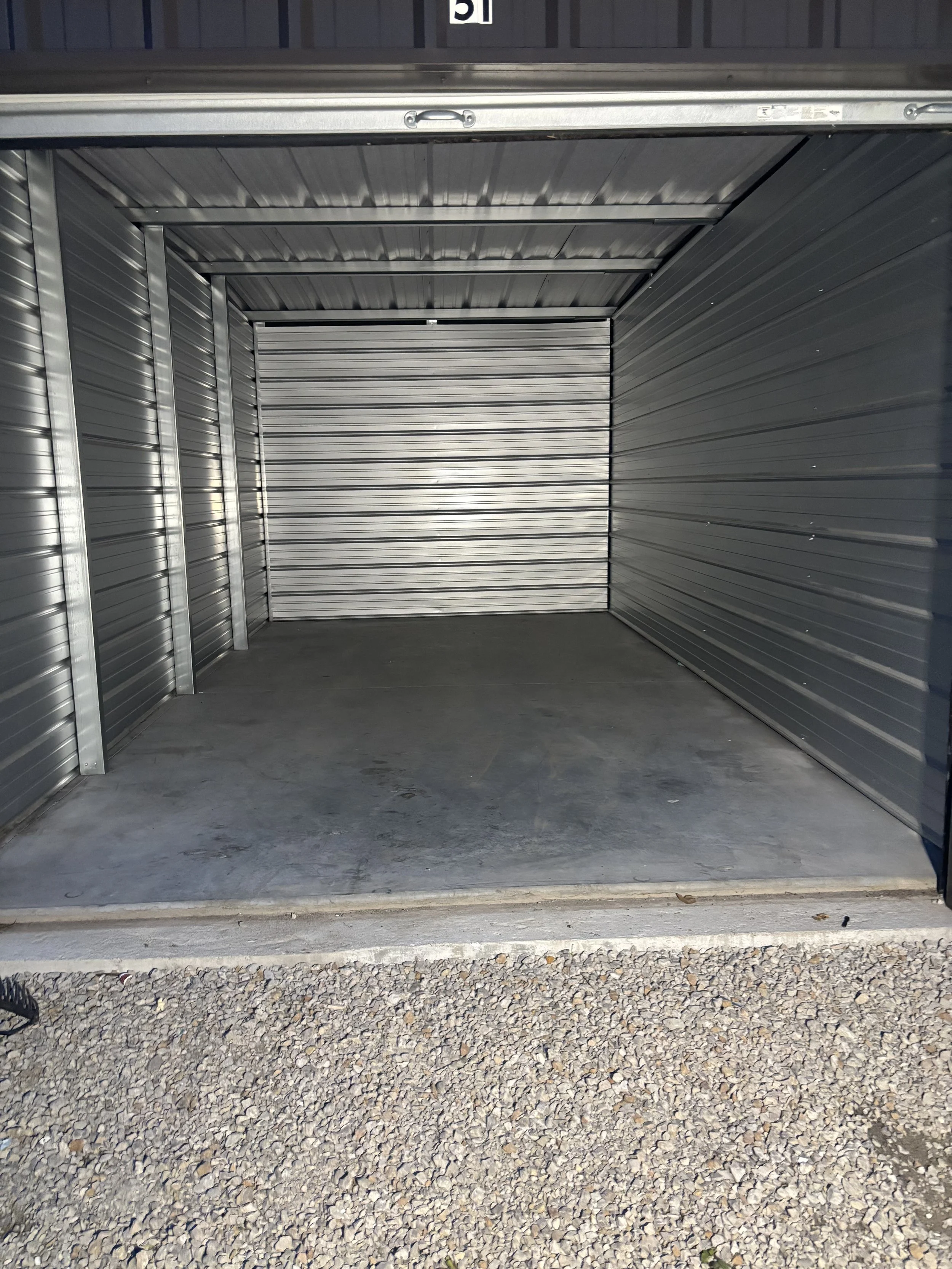 Empty storage unit with open metal rolling doors, concrete floor, and gravel outside.
