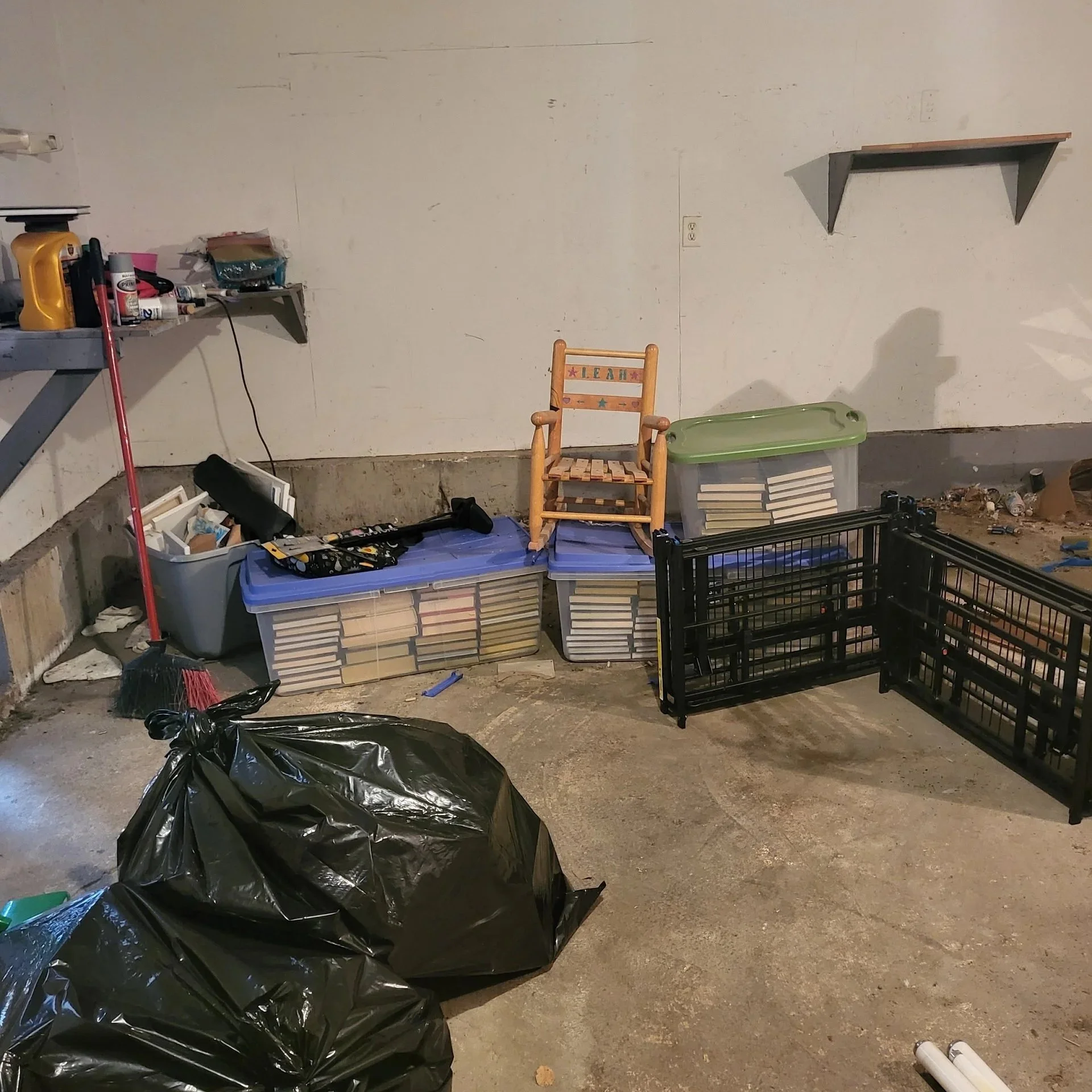 A garage or basement space with clutter including trash bags, plastic storage bins filled with books, a small wooden chair, black plastic crates, a broom, and various tools and items on shelves and the floor.