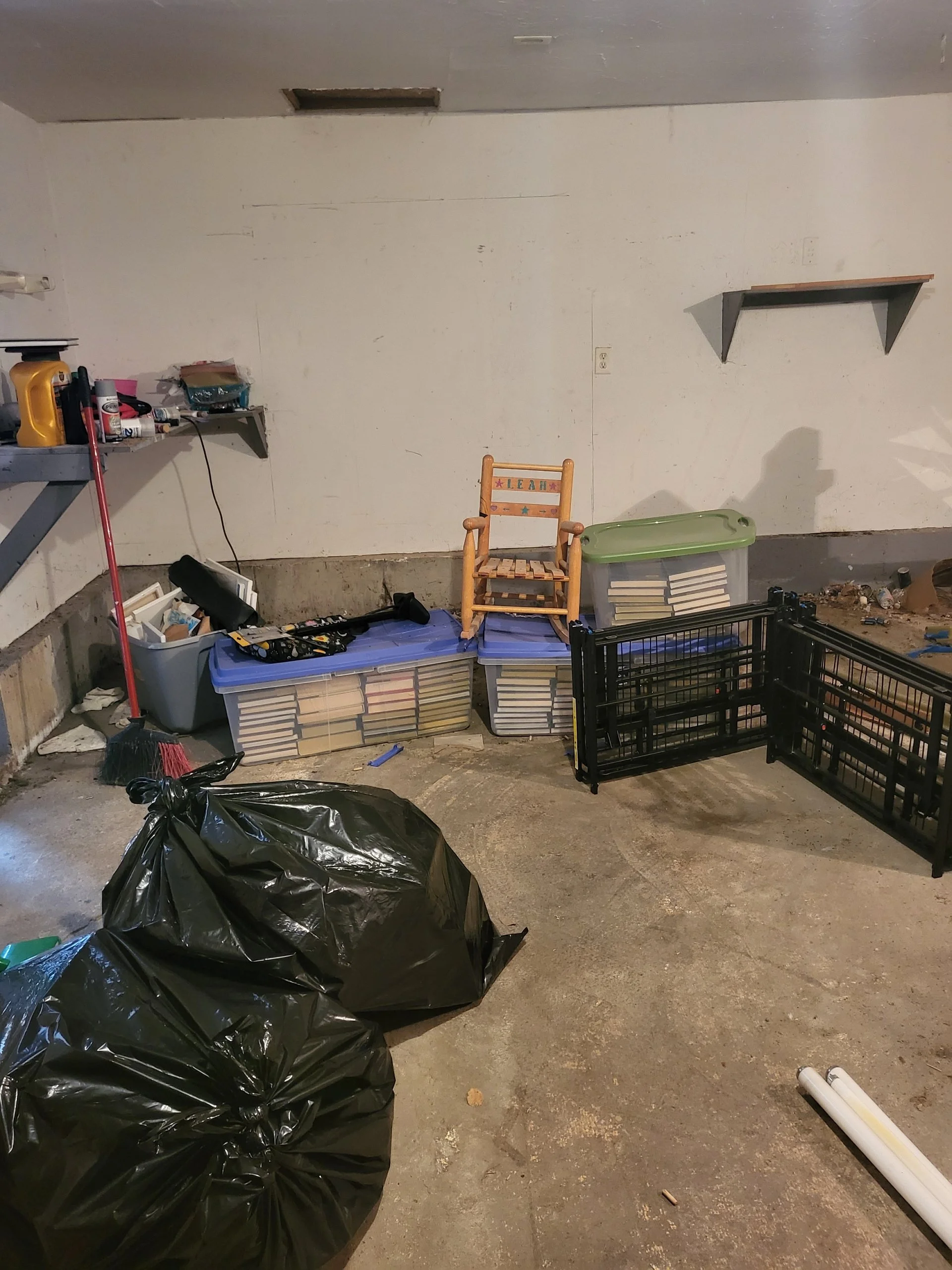 A cluttered garage with two black trash bags on the floor, a plastic storage bin with books, a small wooden chair with the name 'Leah' on it, a shelf with cleaning supplies, and two black plastic crates.