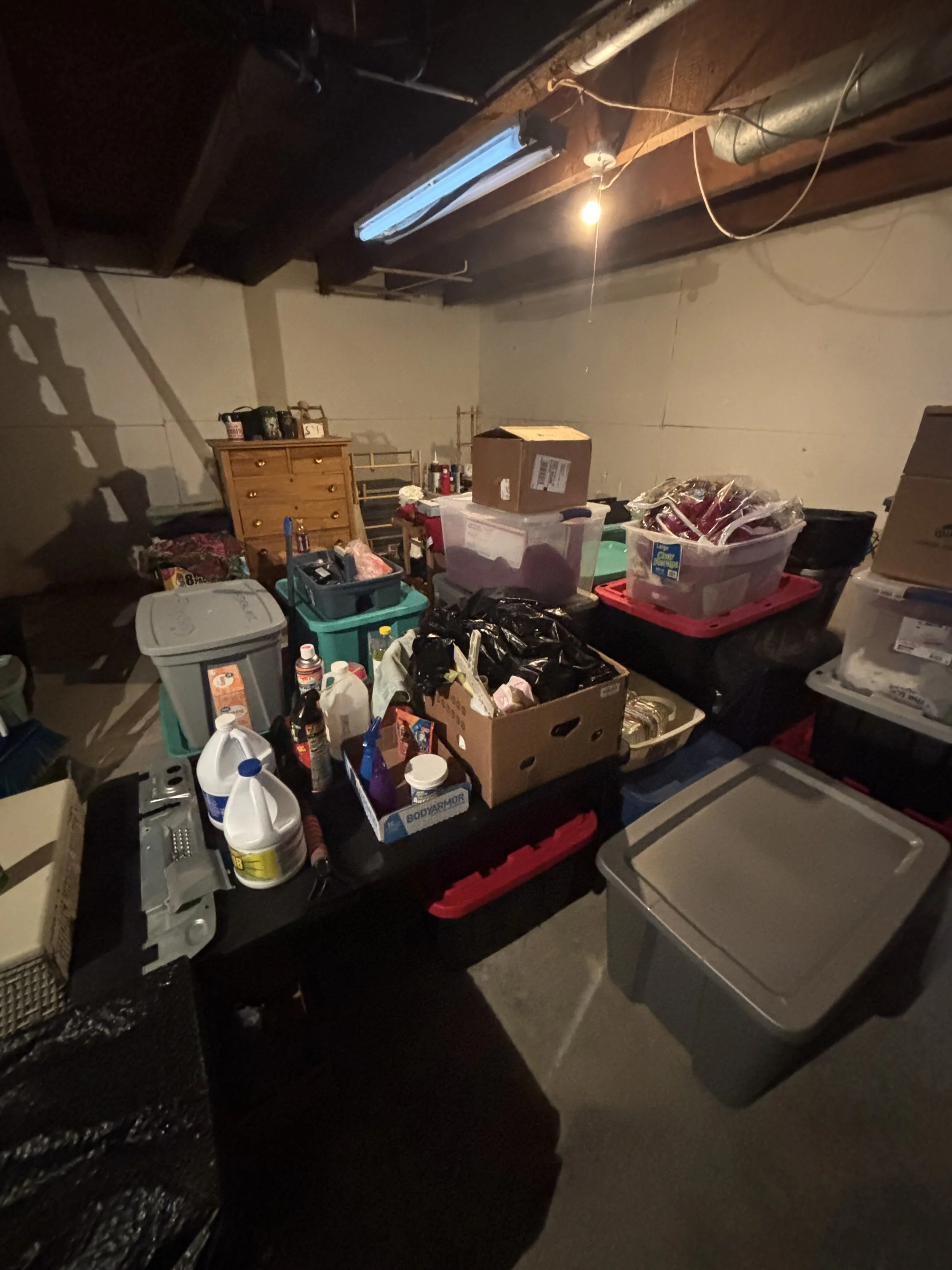 A cluttered basement storage room with various containers, boxes, and miscellaneous items on shelves and tables, with exposed ceiling beams and pipes.