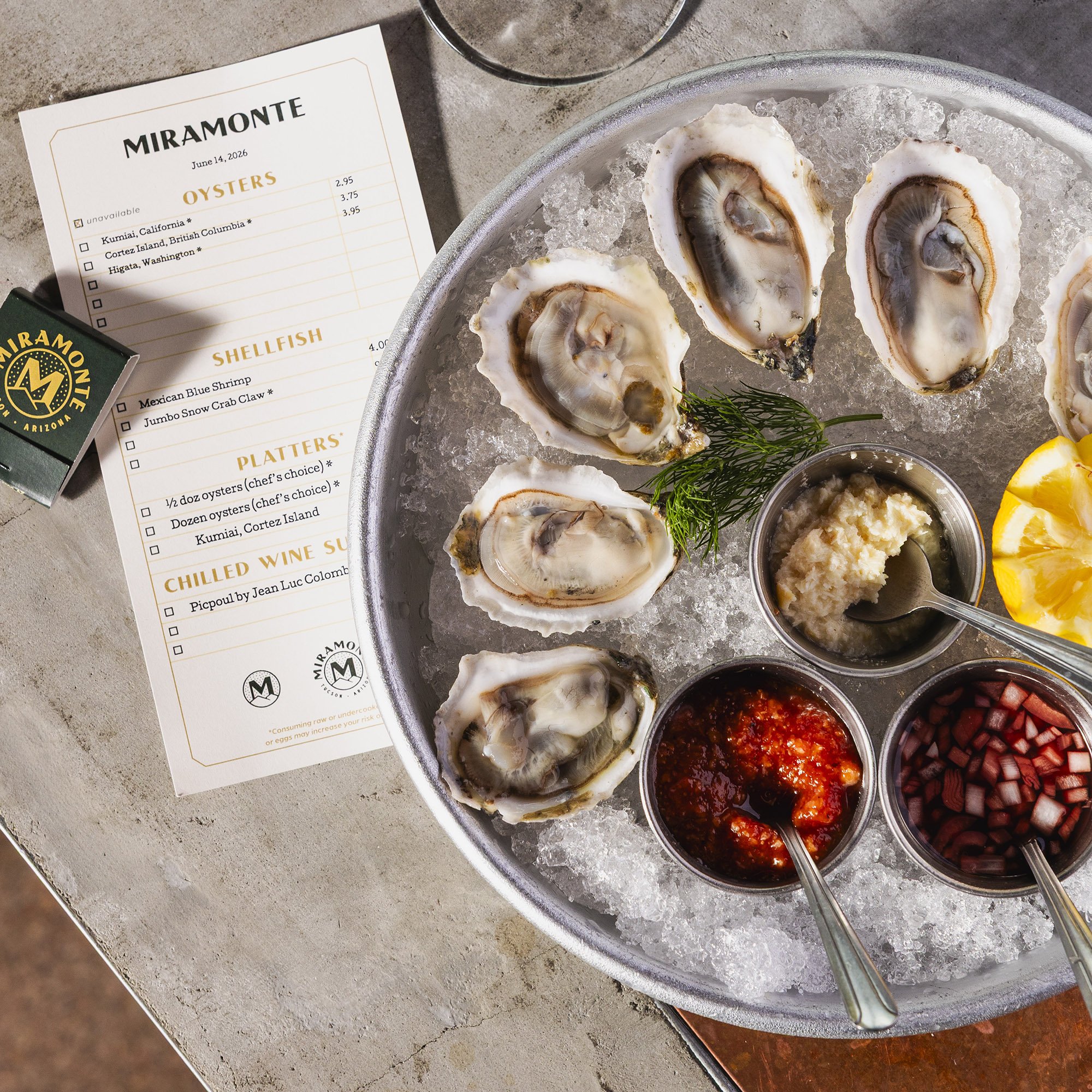 A tray of fresh oysters on ice served with various cocktail sauces, small containers of diced vegetables, and lemon wedges, with a menu card listing oyster options and prices on the side.
