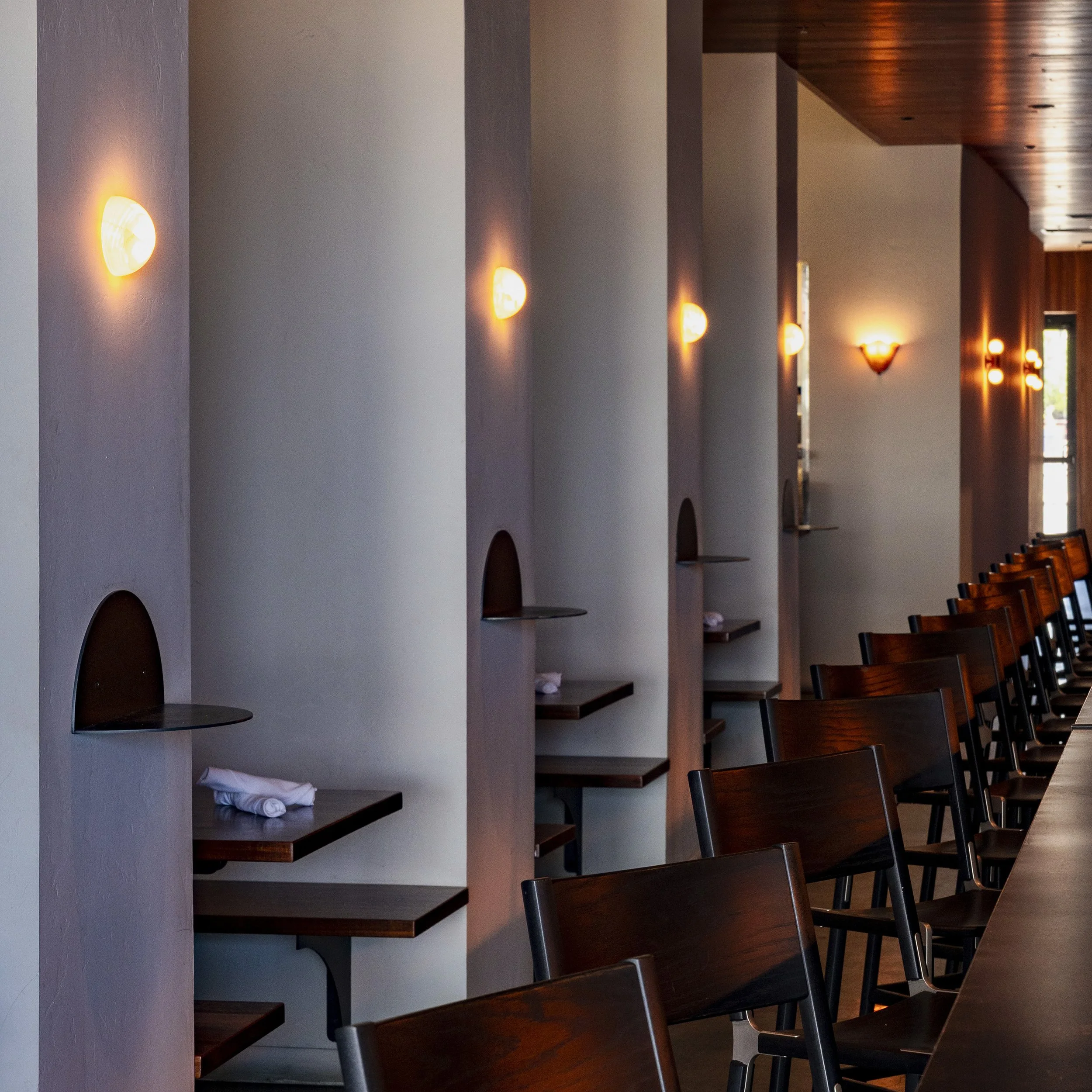 Interior of a modern restaurant or cafe with wall-mounted light fixtures, a row of tables and wooden chairs, and a wooden ceiling.