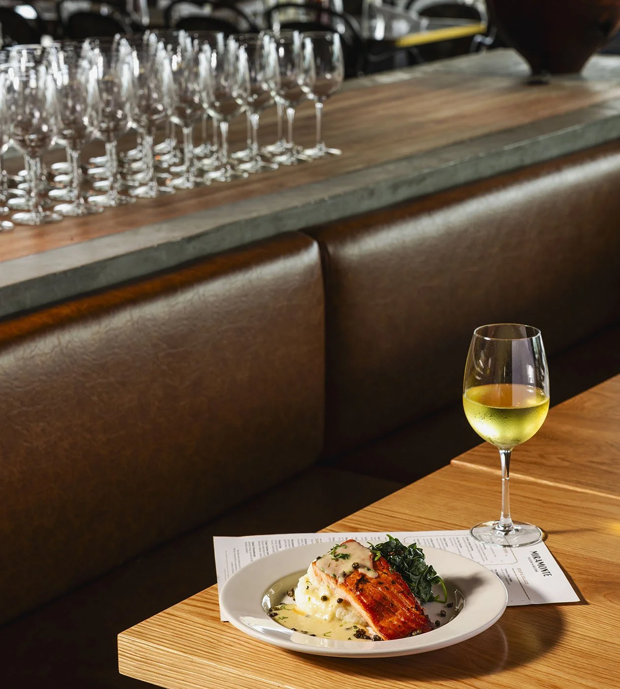 A restaurant table with a plate of salmon on a bed of mashed potatoes, a glass of white wine, a menu, and a glass window with multiple empty wine glasses in the background.