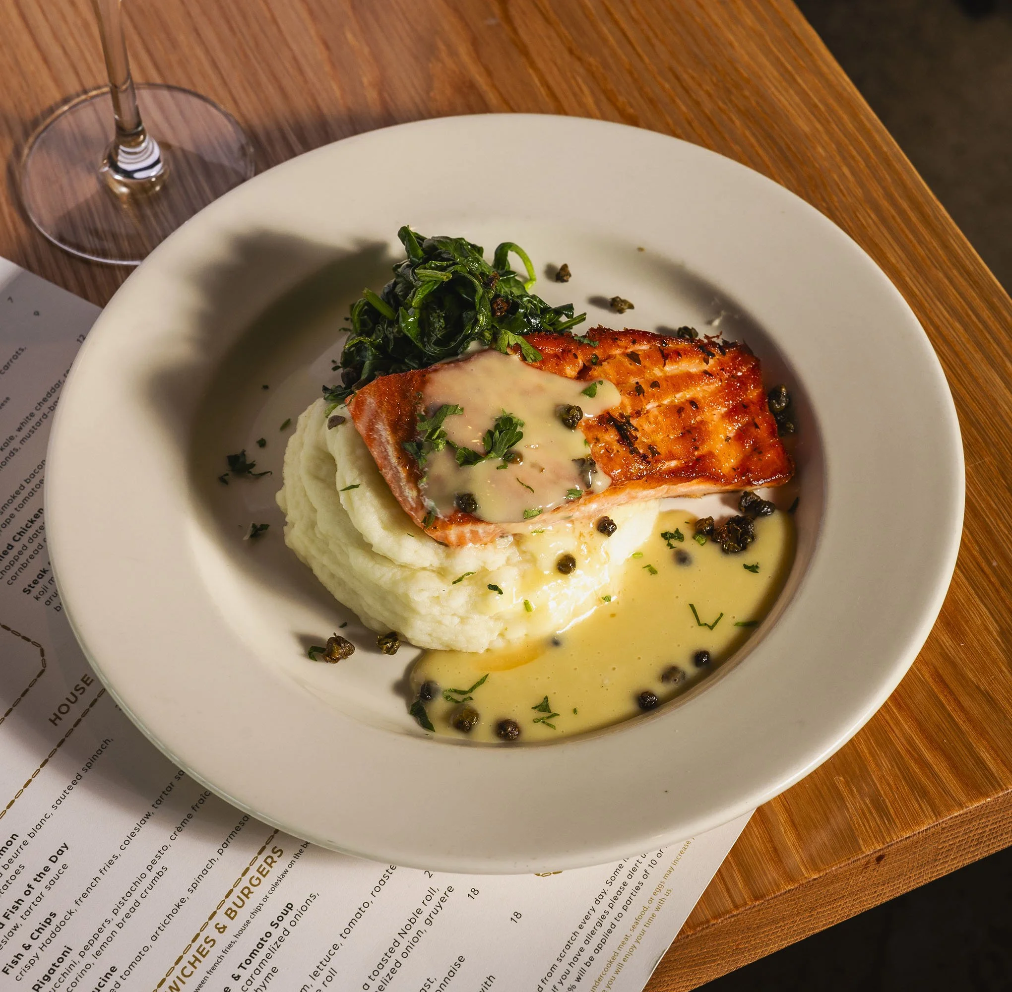 A white plate with grilled salmon topped with creamy sauce, served with a side of cooked spinach, on a wooden table with a glass of wine nearby.