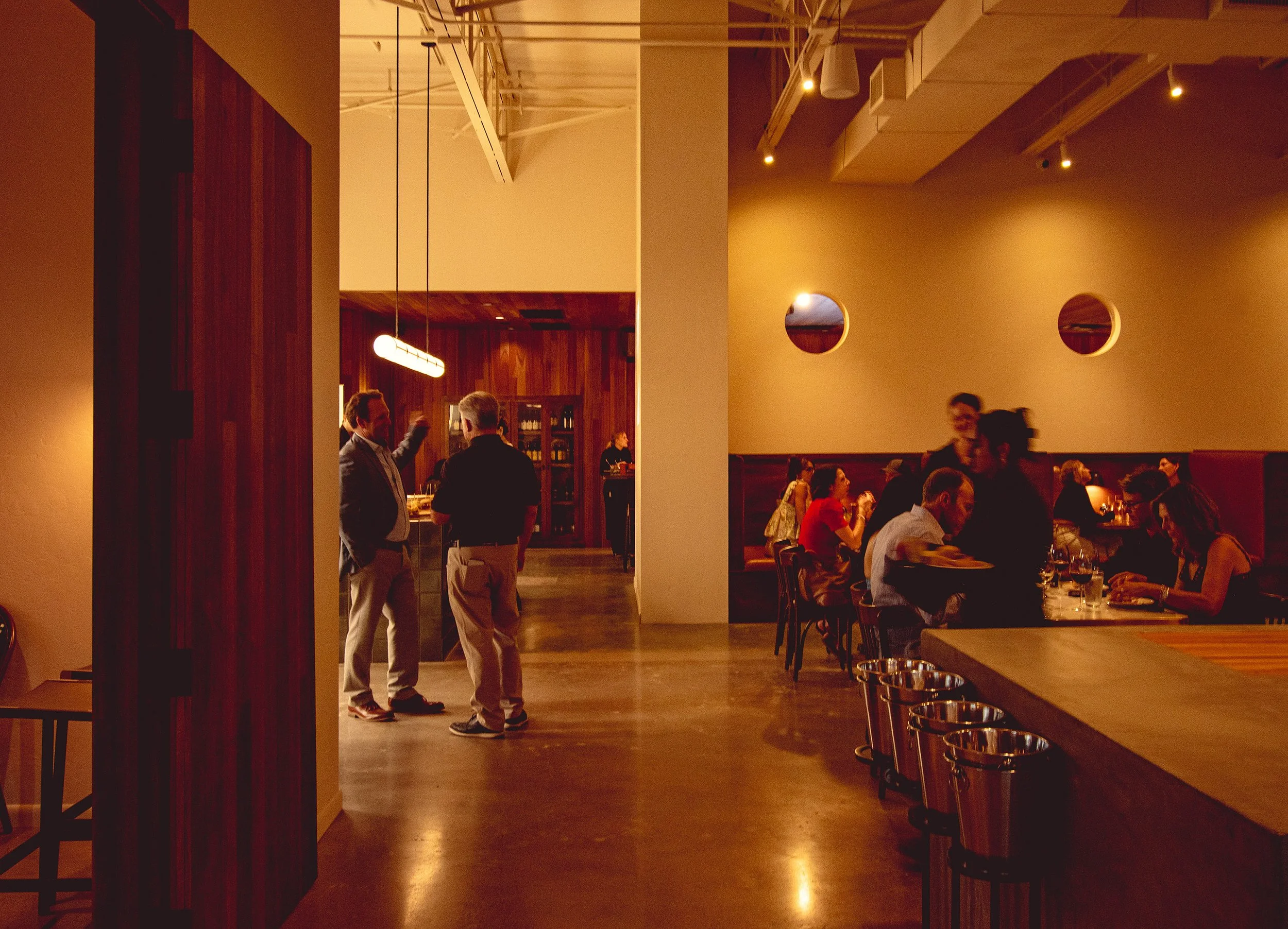 Interior of a restaurant with dim lighting, wooden accents, and several diners seated at tables. There are people standing and conversing near the entrance, and a visible bar area in the background.
