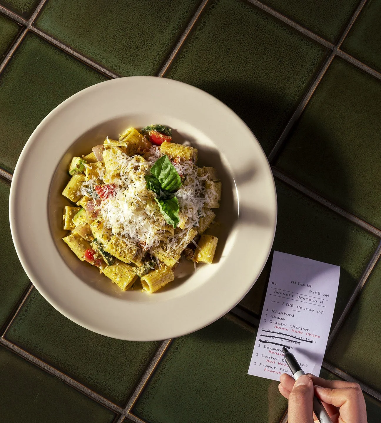 A plate of rigatoni pasta with vegetables and grated cheese, garnished with fresh basil, placed on a tiled green table. A person is writing on a receipt with a black marker.