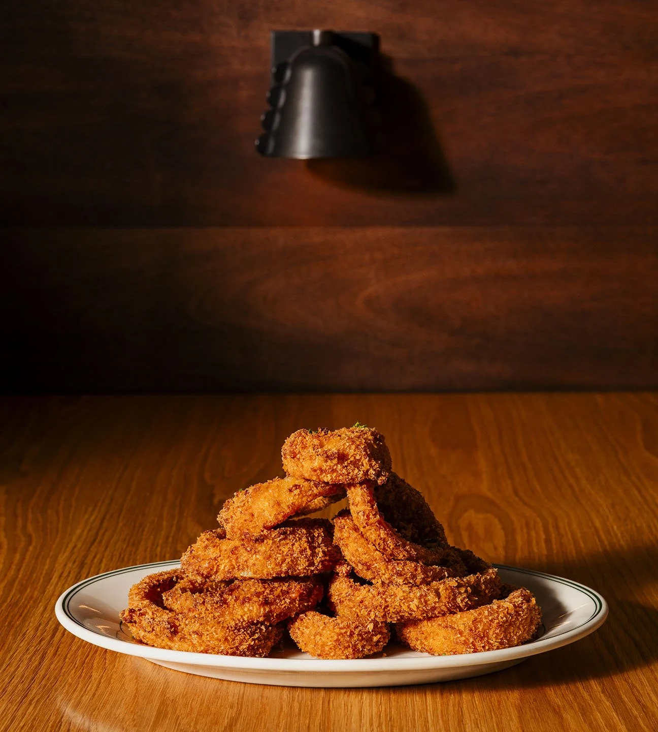 A plate of fried chicken tenders stacked on a wooden table, with a dark wooden background and a focused light above.
