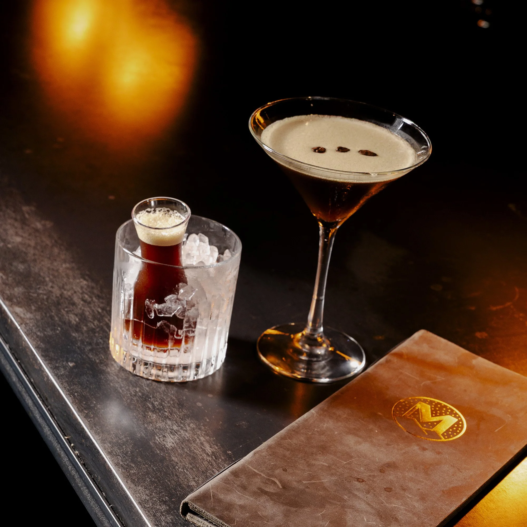 A cocktail in a martini glass garnished with three coffee beans, with a small shot glass and a glass container filled with ice nearby, on a dark bar counter.