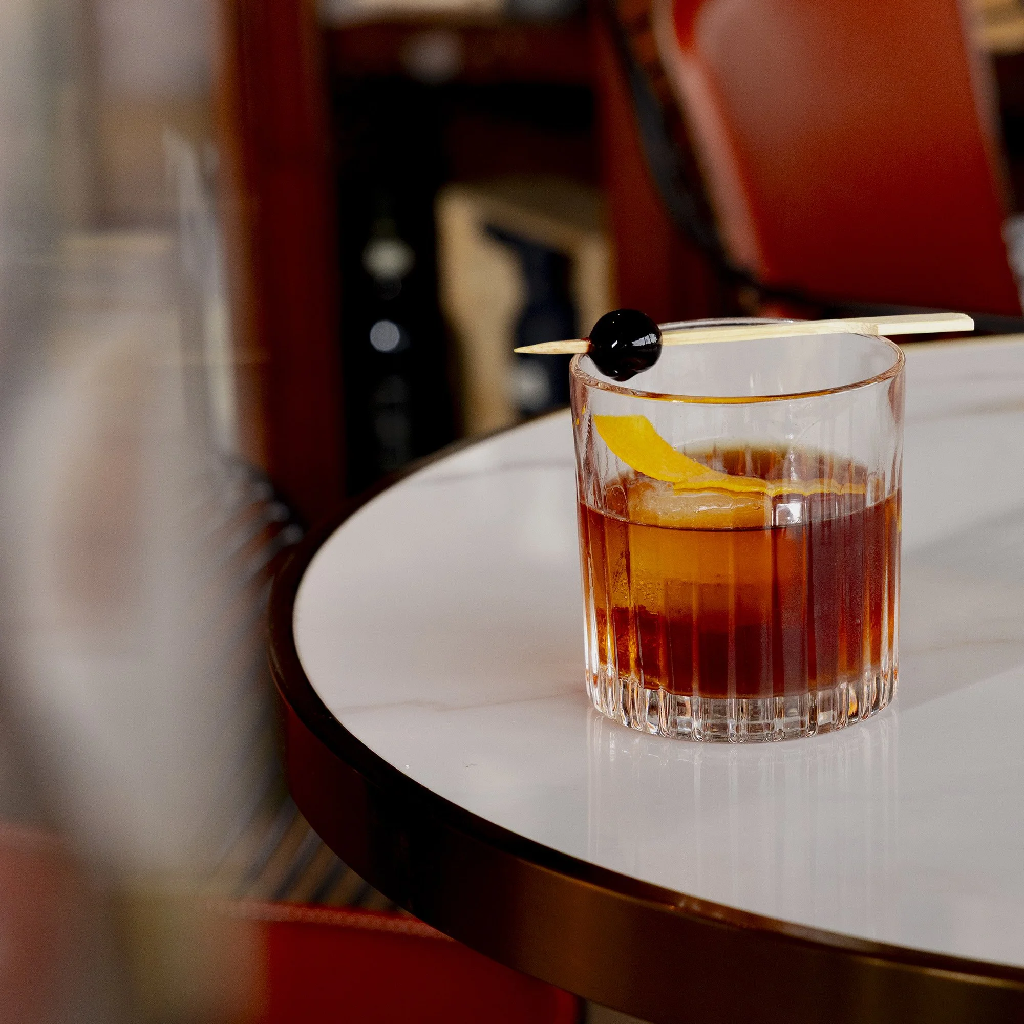 A close-up of a cocktail glass with orange liquid, garnished with a black cherry and a peach slice, resting on a white table.