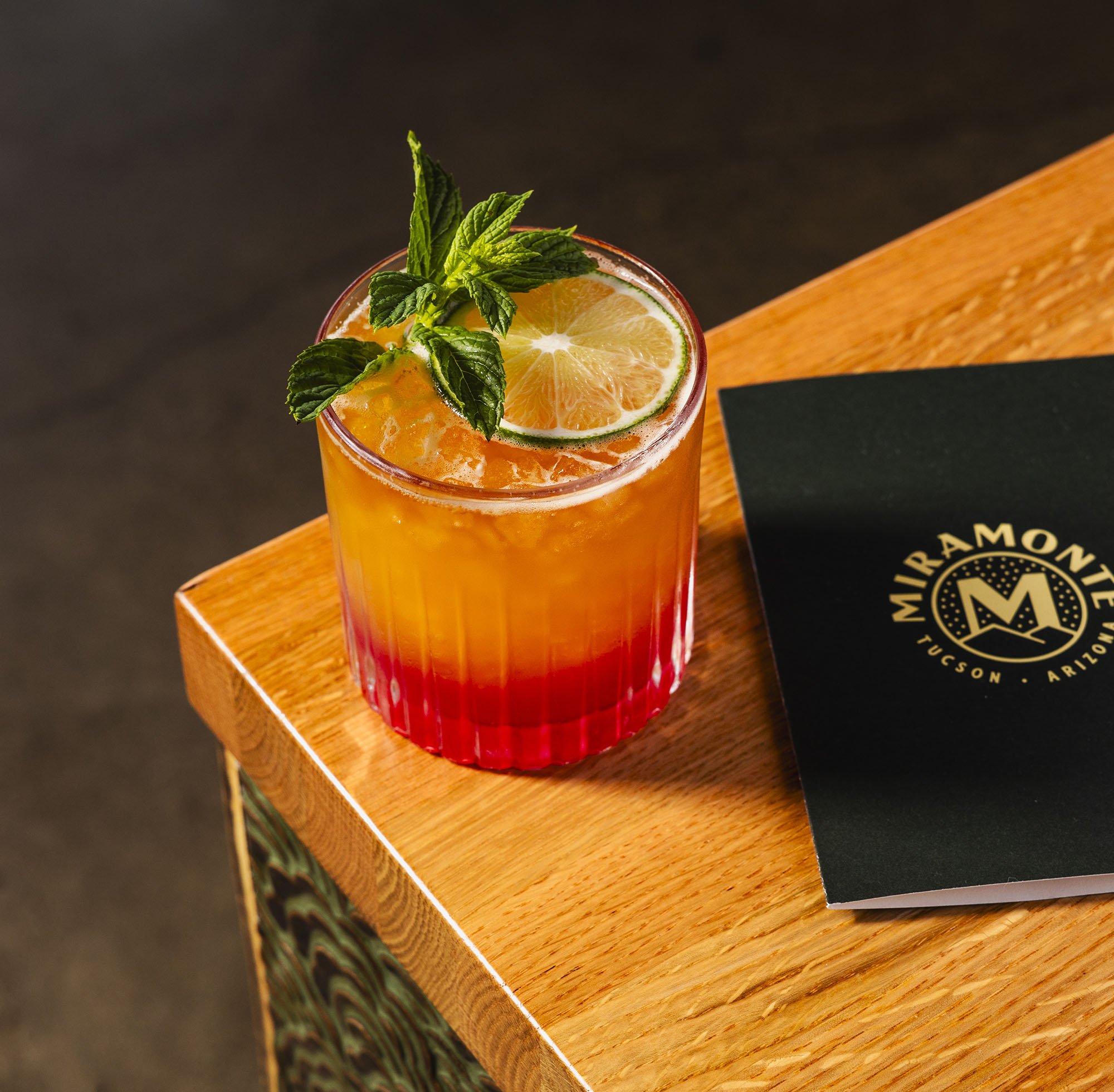 A glass of orange-colored beverage garnished with a sprig of fresh mint and a slice of lemon, placed on a wooden surface.