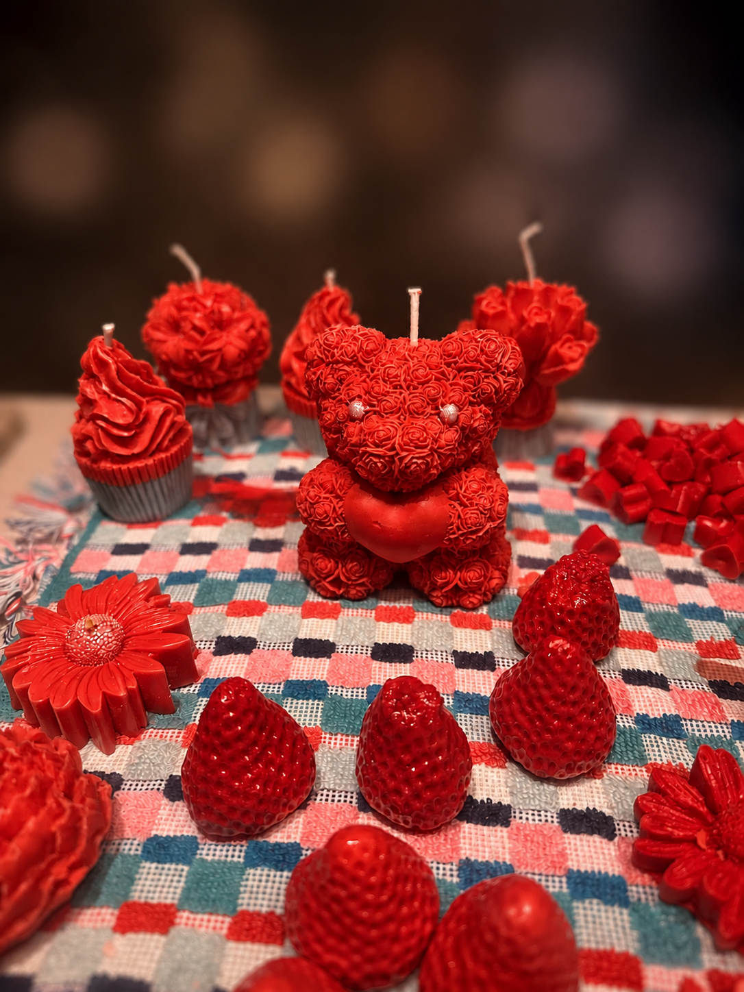 Red teddy bear-shaped candle surrounded by strawberries, cupcakes with red frosting, and decorative flowers on a checkered tablecloth.