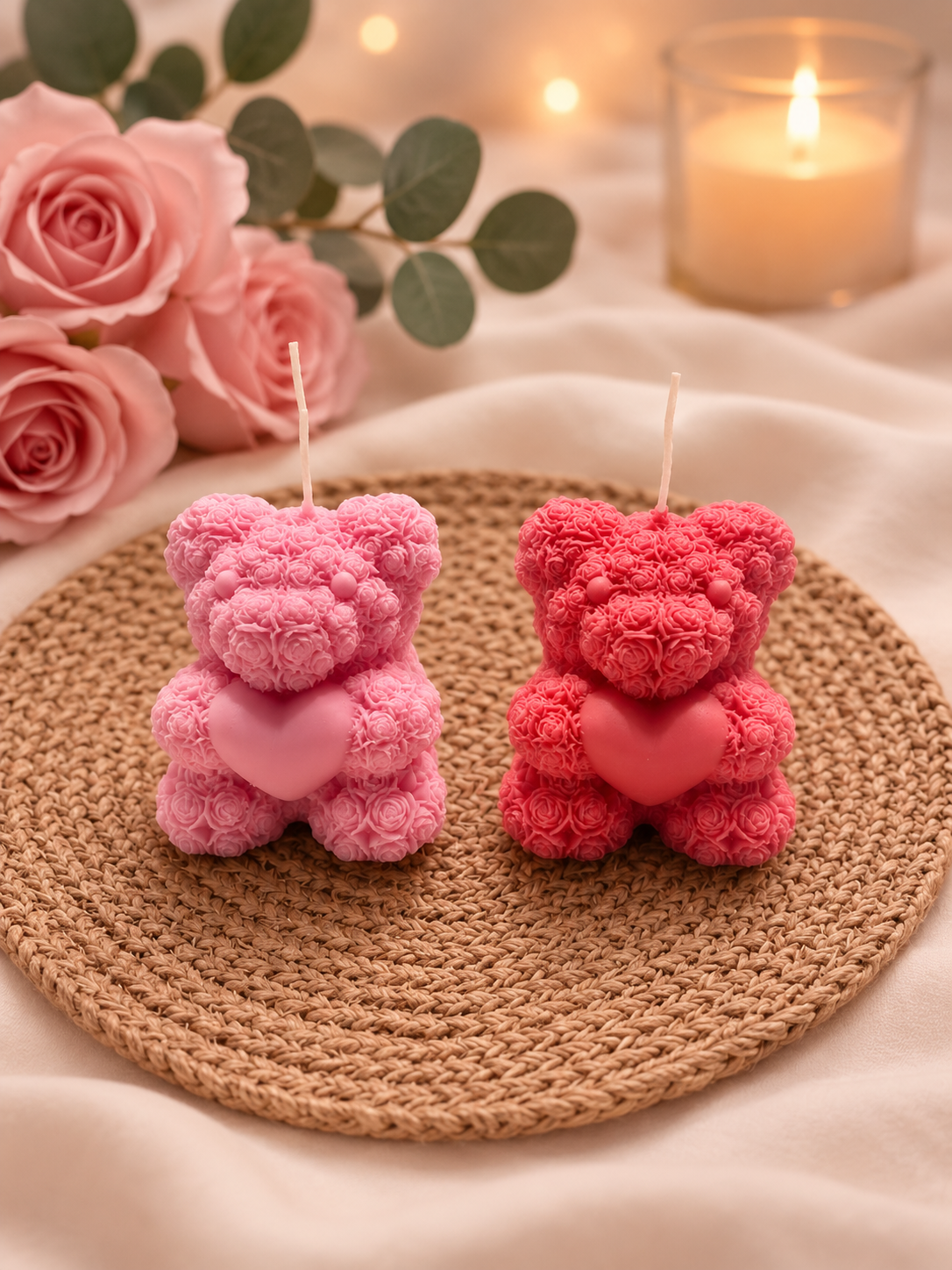 Pink and red teddy bear-shaped candles with heart accents on a woven round placemat, with pink roses, eucalyptus leaves, and a lit candle in the background.