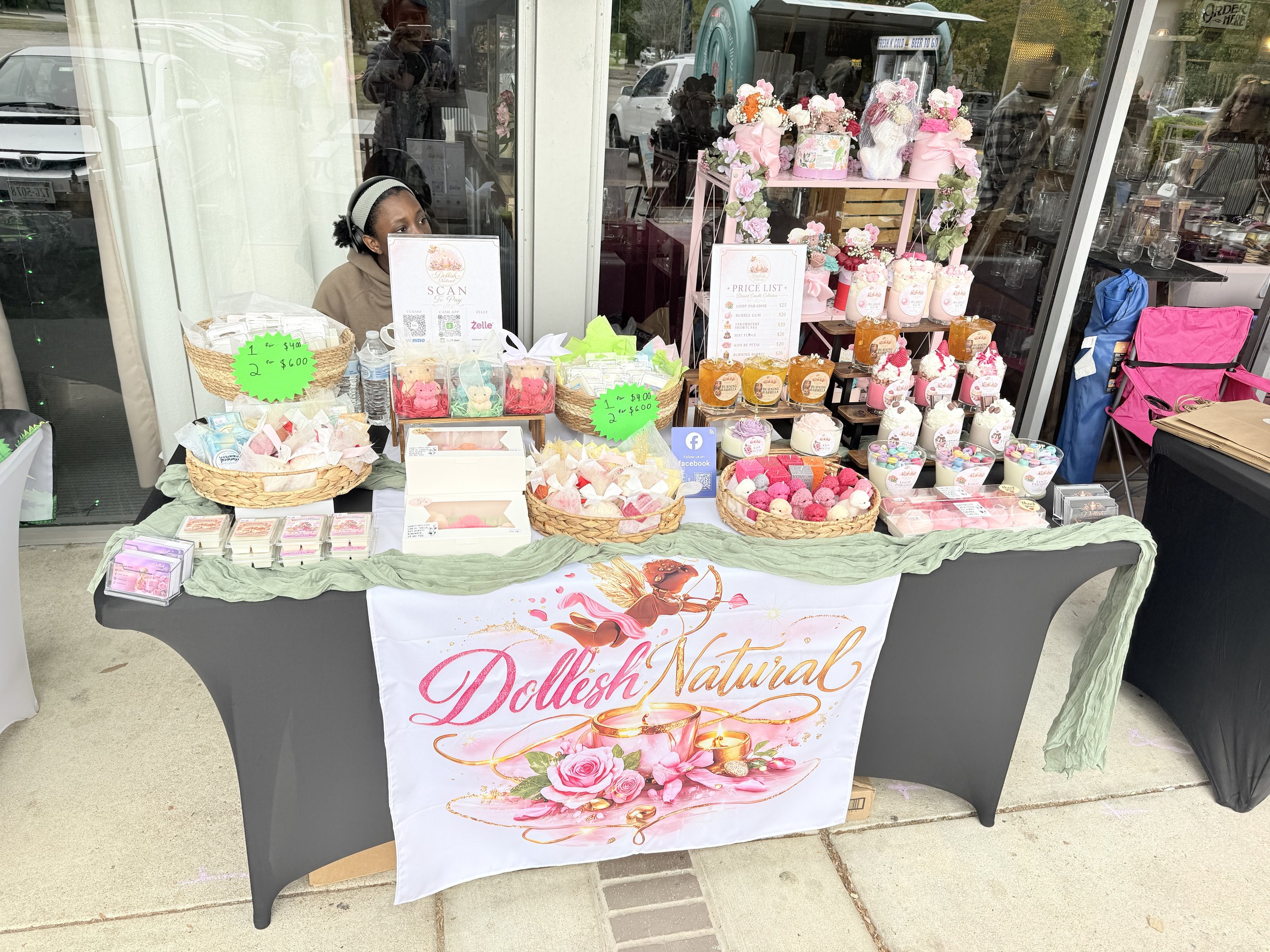 A table with pink flowers, bath bombs, and colorful bath products, displayed in a store window with a sign promoting natural dolls and a price list. A person with headphones is sitting behind the table.