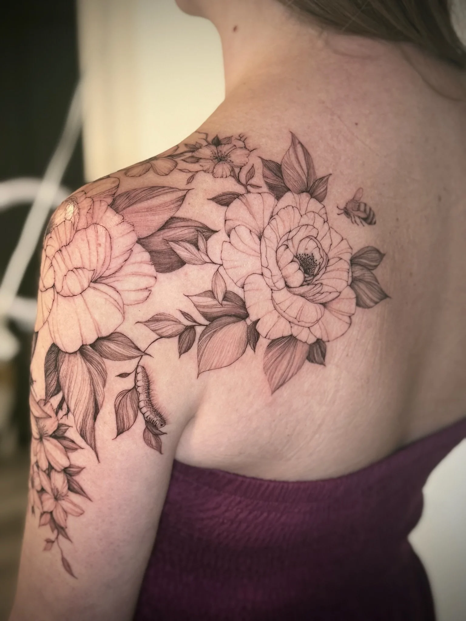 Close-up of a person's shoulder and upper arm covered with a detailed floral, fine-line tattoo design, featuring flowers, leaves, and a bee, with a maroon sleeveless top.