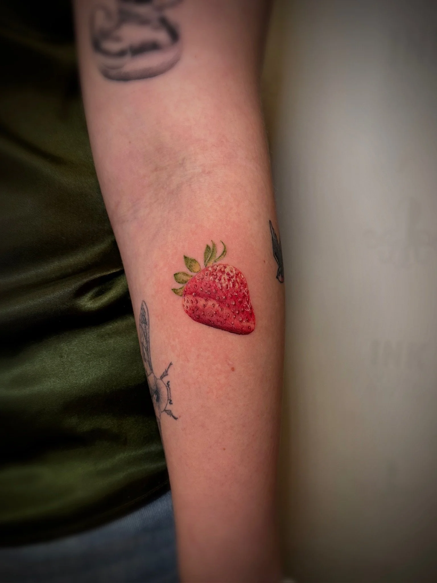 Close-up of a person's forearm with a tattoo of a fine-line strawberry and some other smaller tattoos nearby.