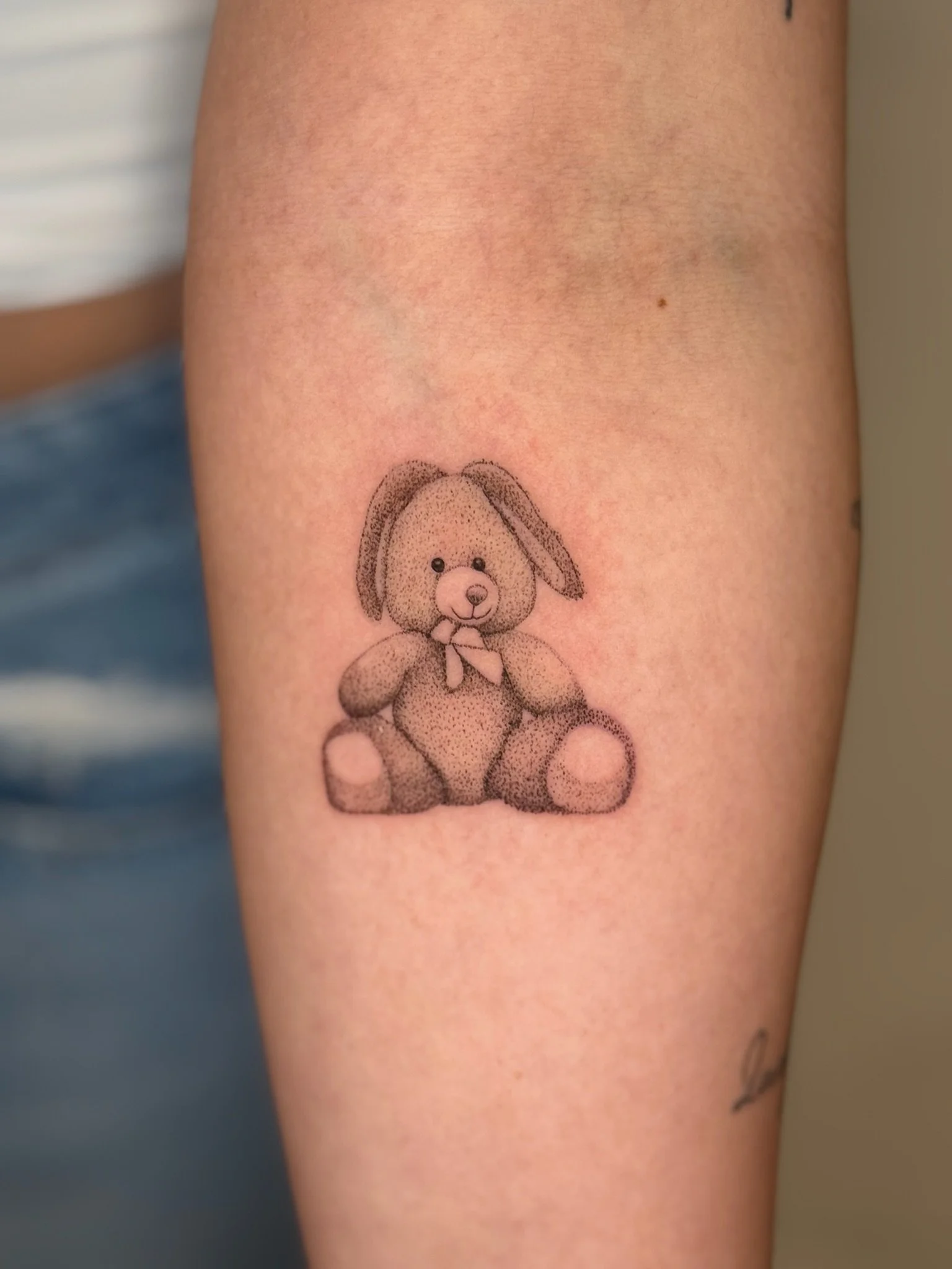 Tattoo of a teddy bear with a bowtie on a person's upper arm.