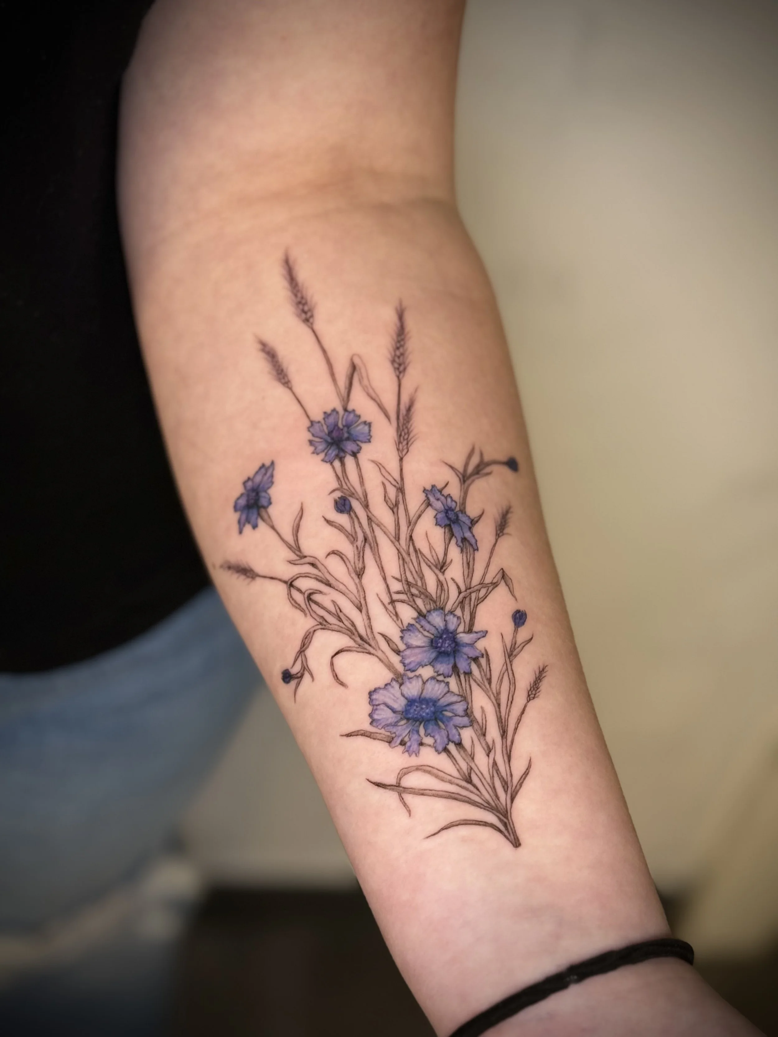 A tattoo of purple flowers with green leaves and brown stems on a person's forearm.