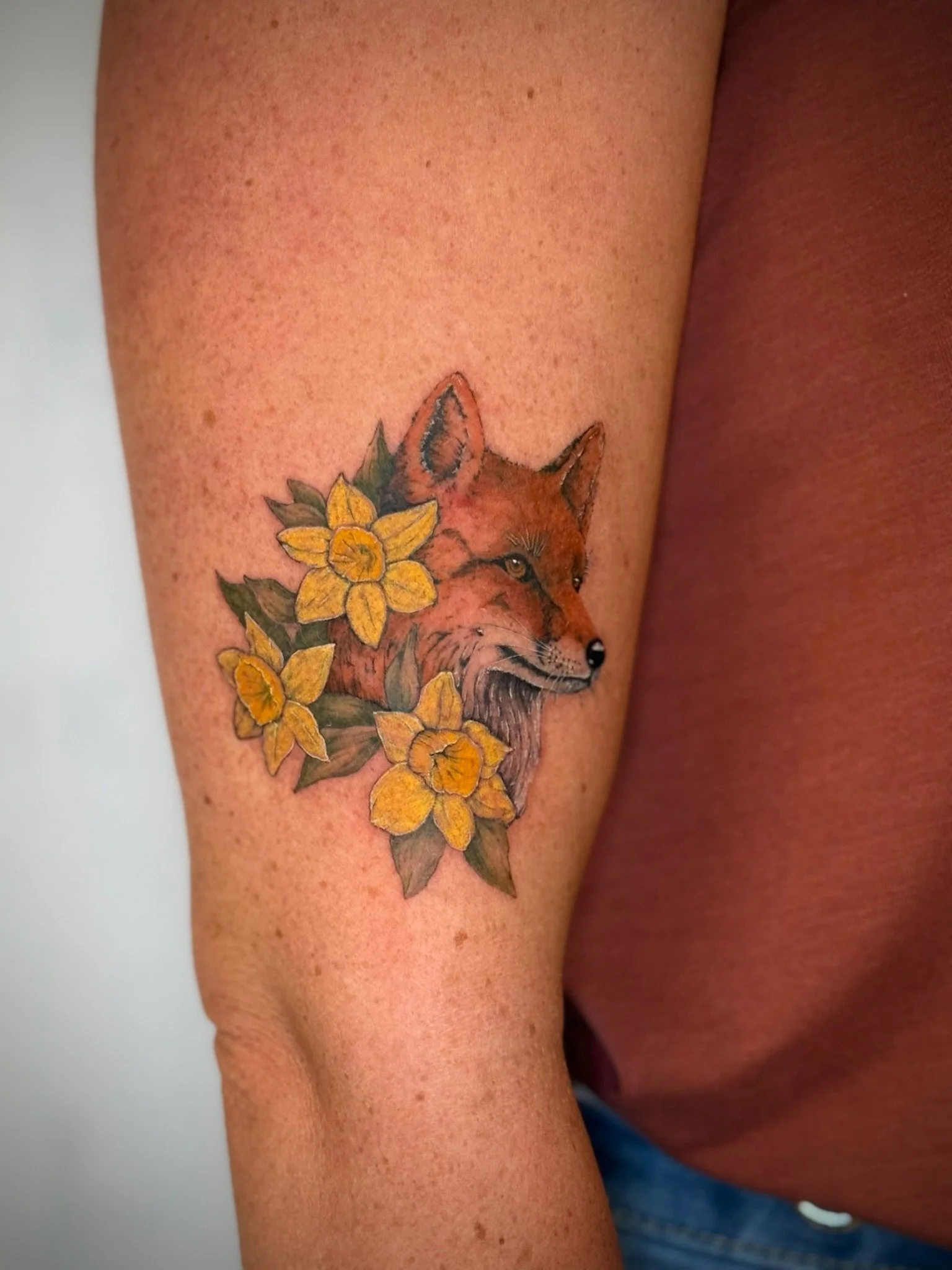 A tattoo of a fox with orange and brown fur, surrounded by yellow flowers with green leaves on a person's upper arm.