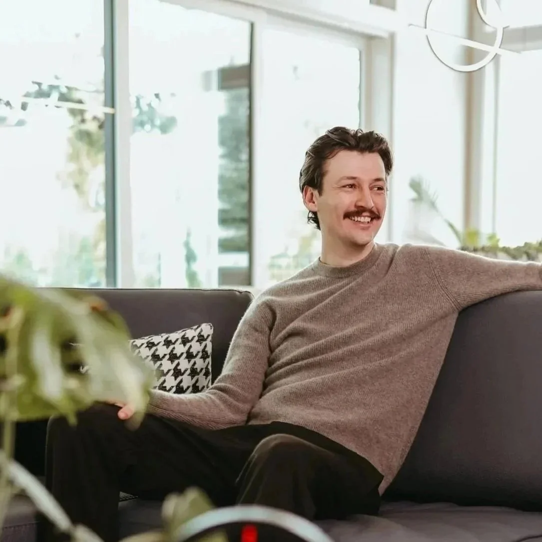 Gage Mansfield, a fine-line tattoo artist, is sitting on a dark gray sofa, smiling and relaxing in a bright room with large windows and houseplants.