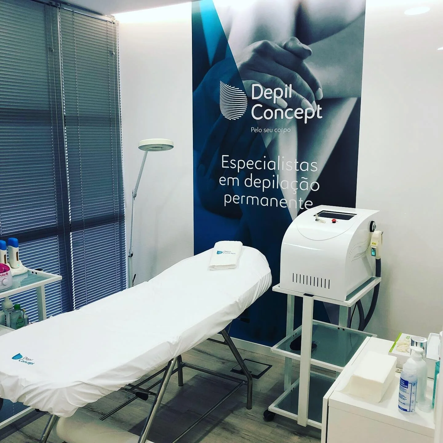 Sala de estética com mesa de tratamento coberta por lençol branco, máquinas de depilação a laser, e uma parede com o logo e textos de divulgação do serviço de depilação permanente. O ambiente é limpo e organizado, com cortinas de janela e iluminação adequada.