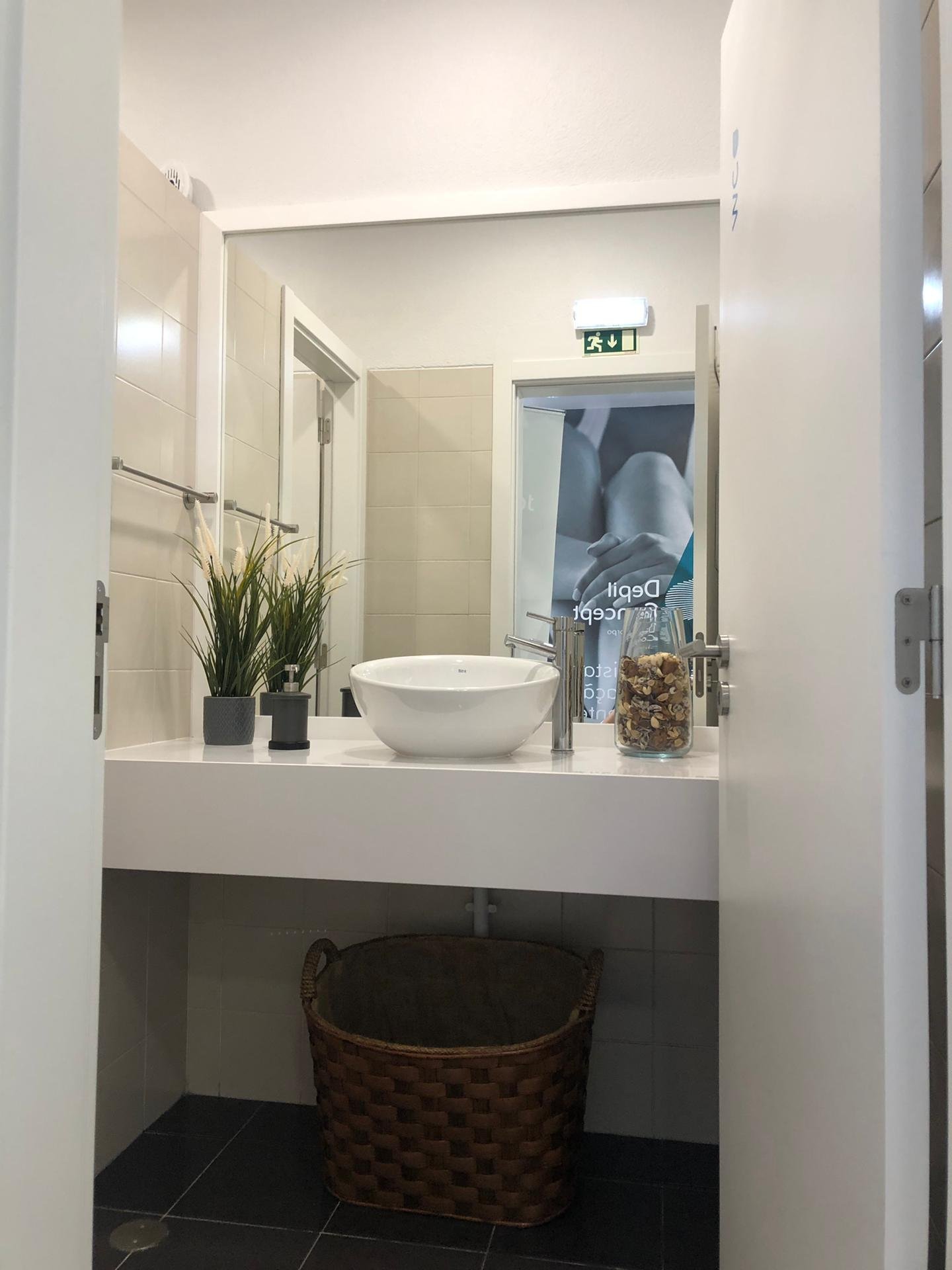Banheiro com pia de bancada branca, espelho grande, vaso de vaso, plantas decorativas, recipientes de acessórios e uma cesta de fibra na parte inferior, com porta de correr ao fundo e uma placa de emergência acima da porta.