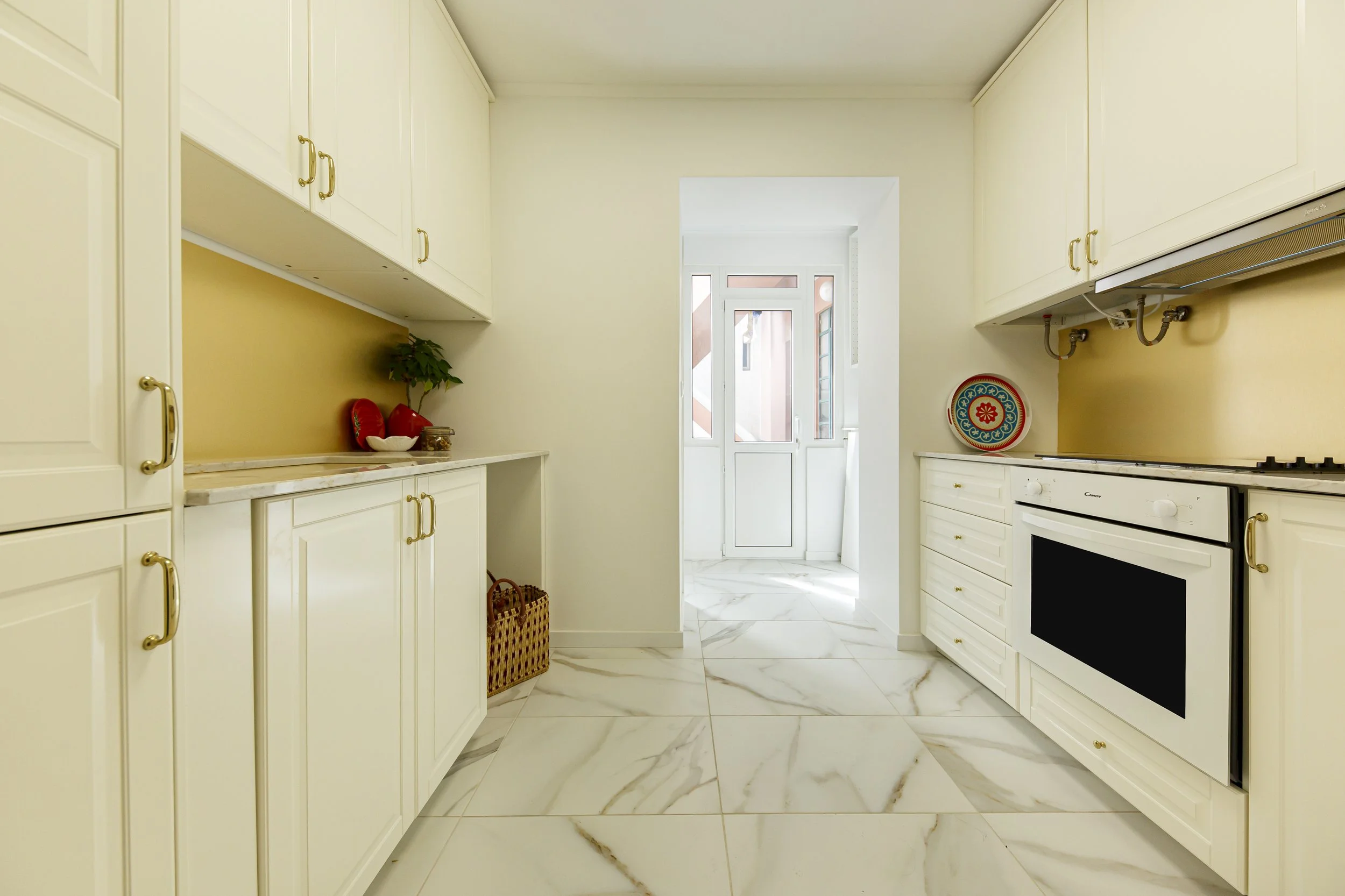 Cozinha com armários brancos, piso de mármore, e uma porta de vidro ao fundo que leva a uma área externa.