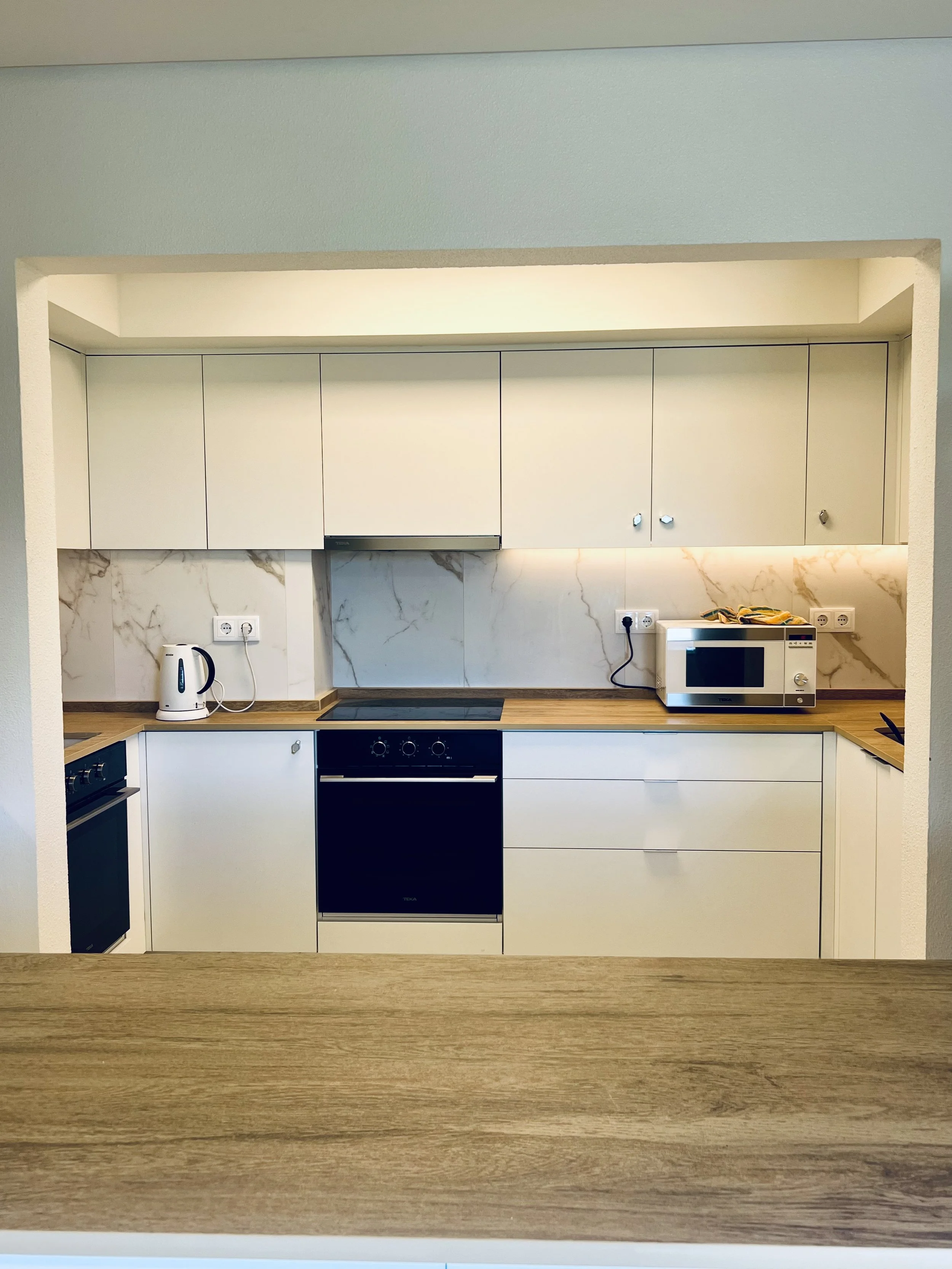 Cozinha moderna com armários brancos, bancada de madeira, forno, micro-ondas, chaleira elétrica e cooktop, decorada com bancada de mármore
