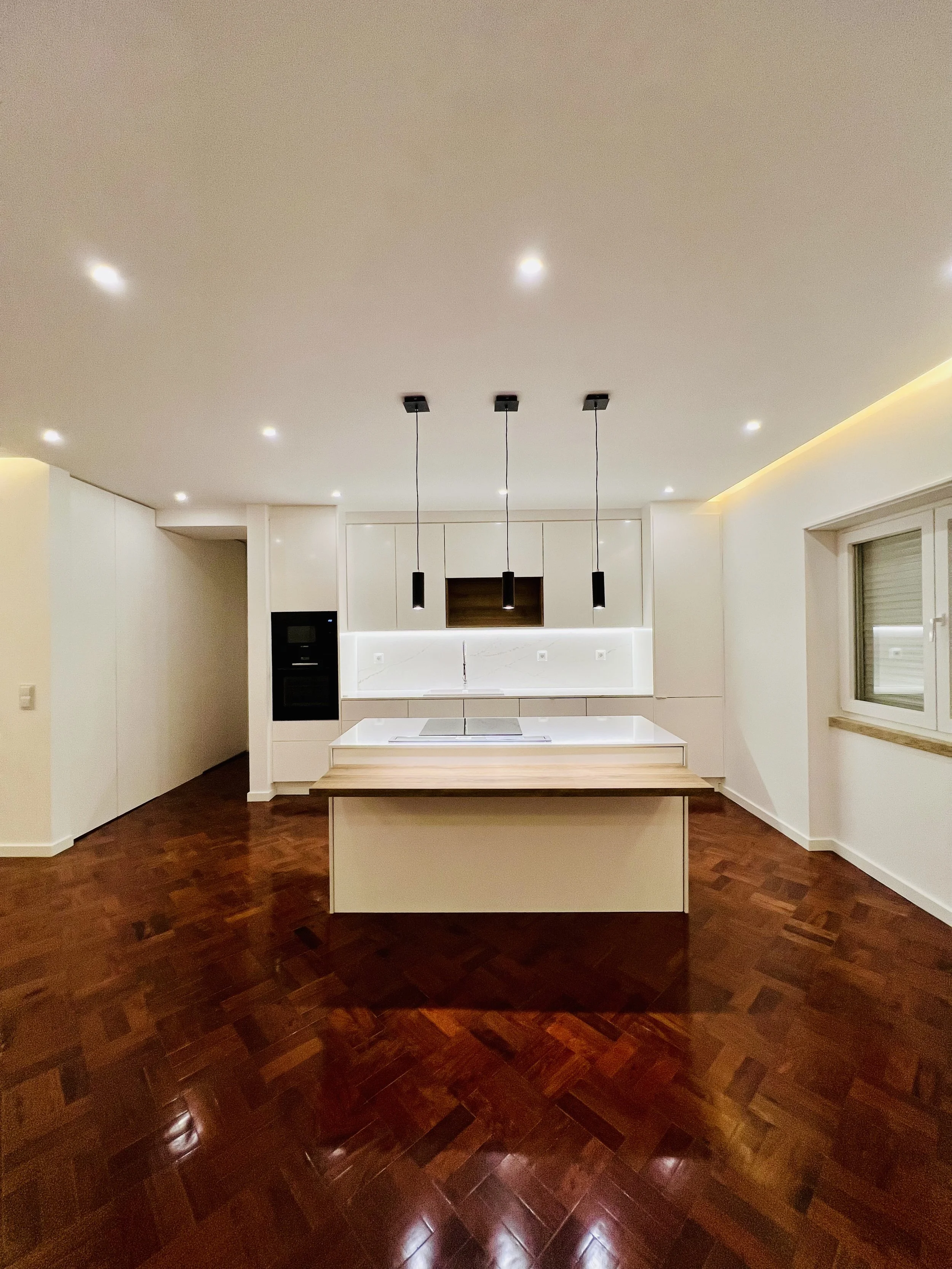 Cozinha moderna com armários brancos, ilha central com bancada de mármore, prateleira de madeira, iluminação embutida e três luminárias pendentes pretas. Piso de madeira escura e janela ao lado com vista para fora.