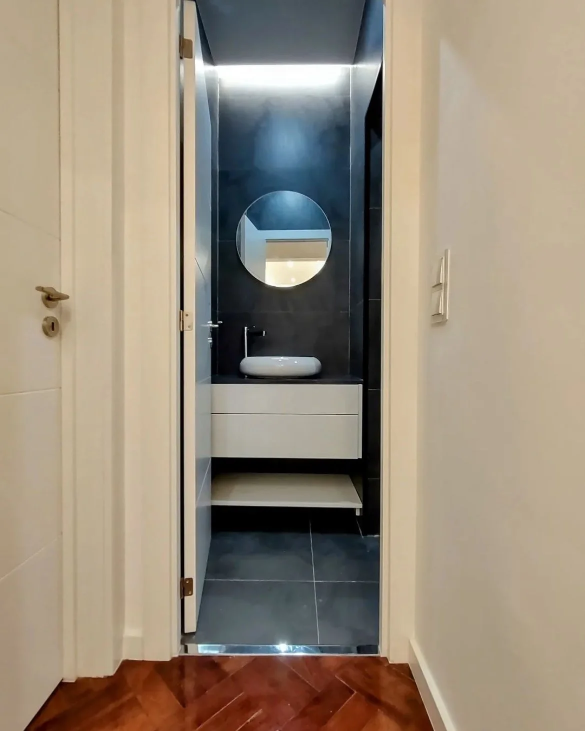 Banheiro pequeno com parede preta, espelho redondo, pias branca com bancada preta, teto com iluminação e porta de correr.