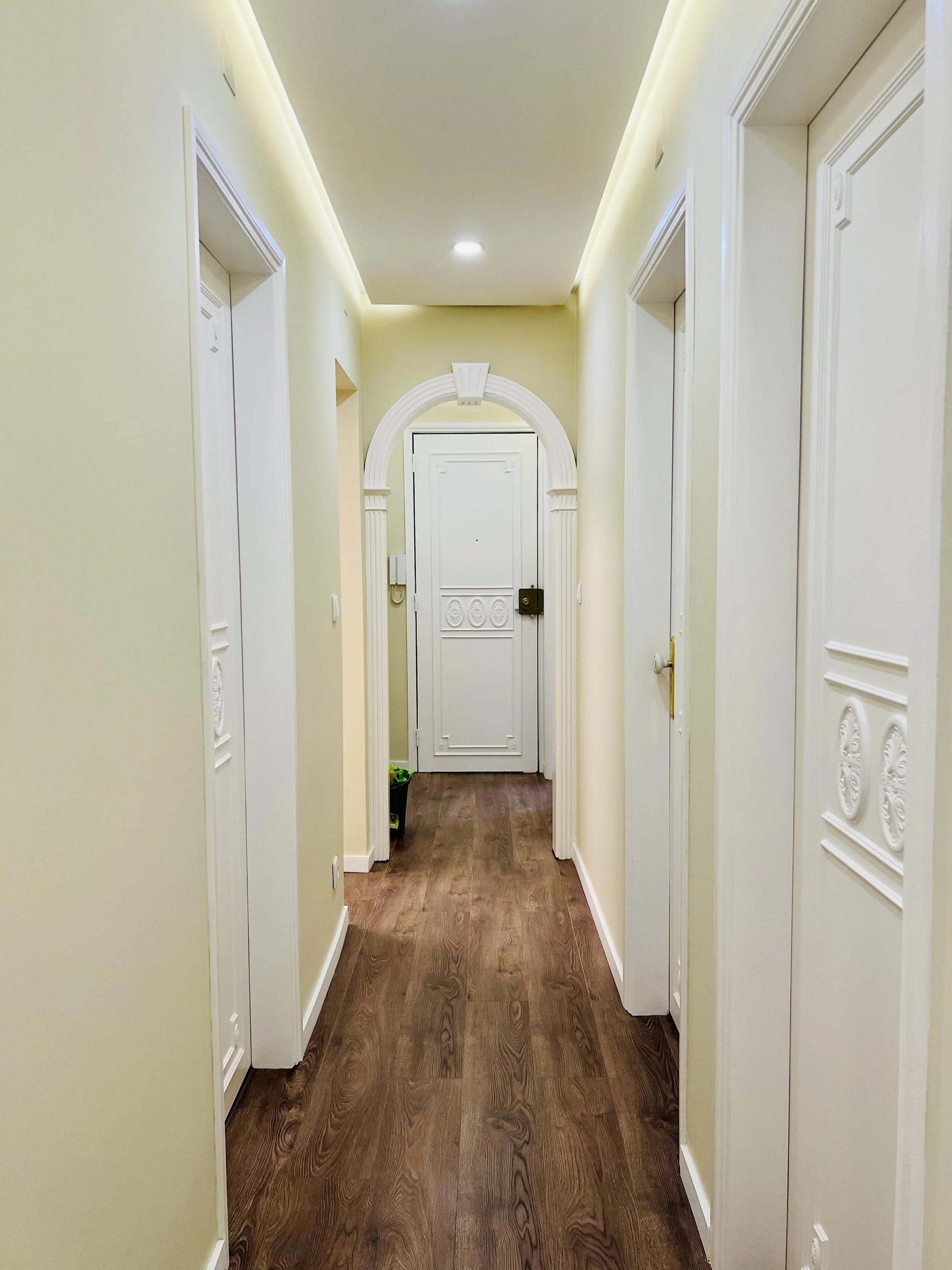 Corredor de apartamento com paredes claras, portas brancas decoradas e piso de madeira, iluminado por luzes embutidas no teto.