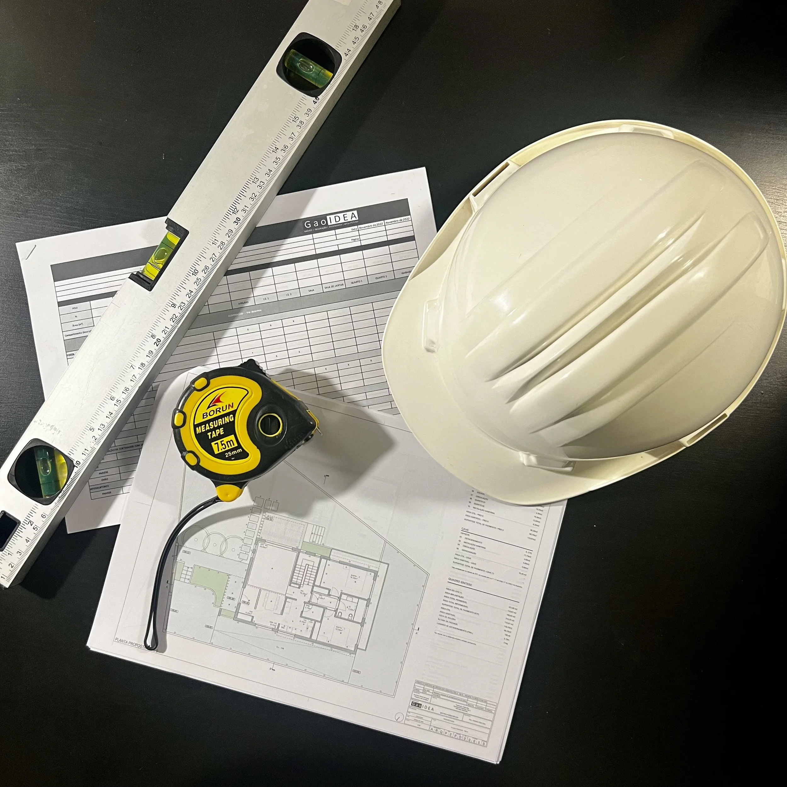 Equipamentos de construção, incluindo uma tábua de nível, uma fita métrica, um capacete de segurança e plantas arquitetônicas sobre uma superfície escura.