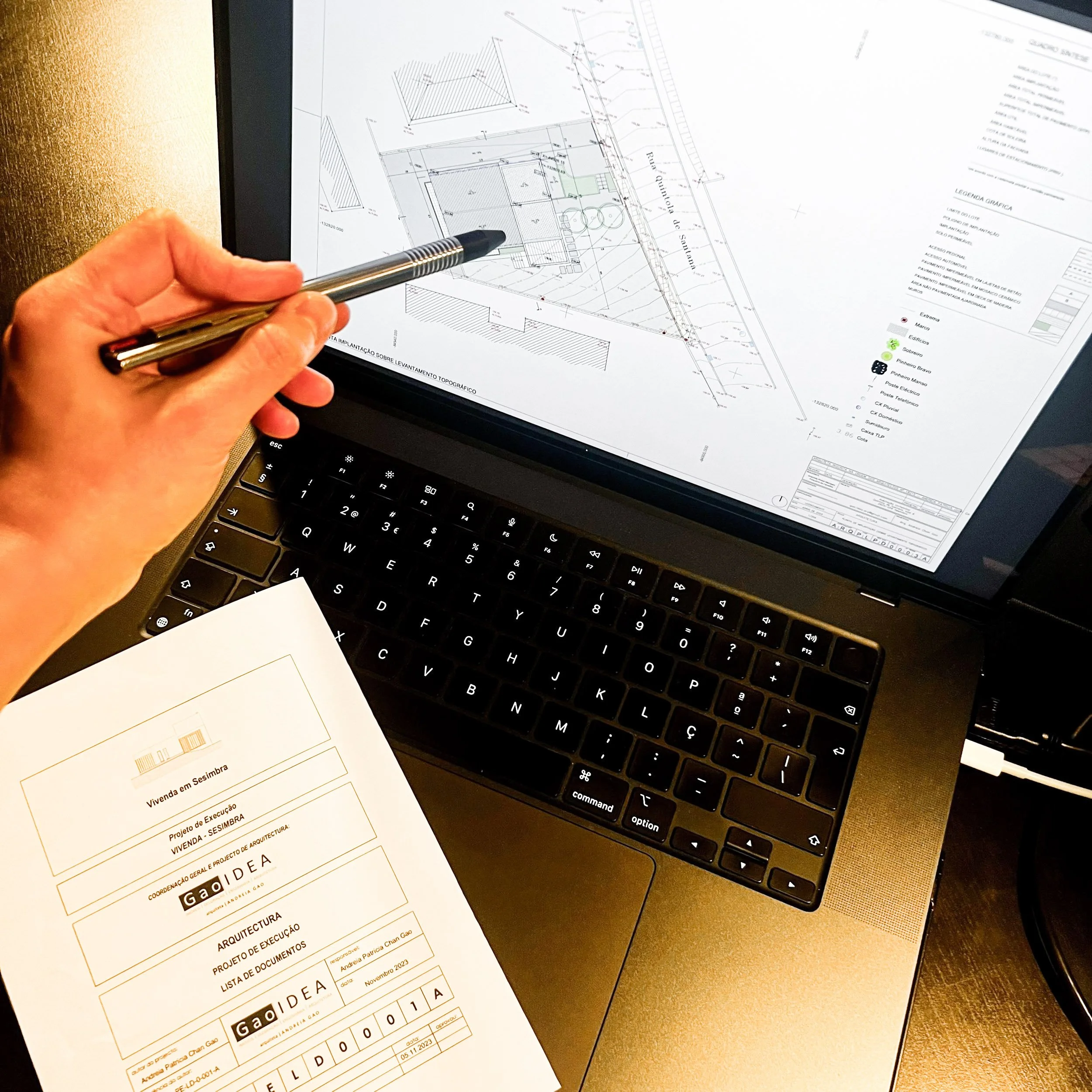Pessoa analisando um projeto de engenharia digital em um computador portátil, com uma folha de papel contendo detalhes do projeto ao lado.