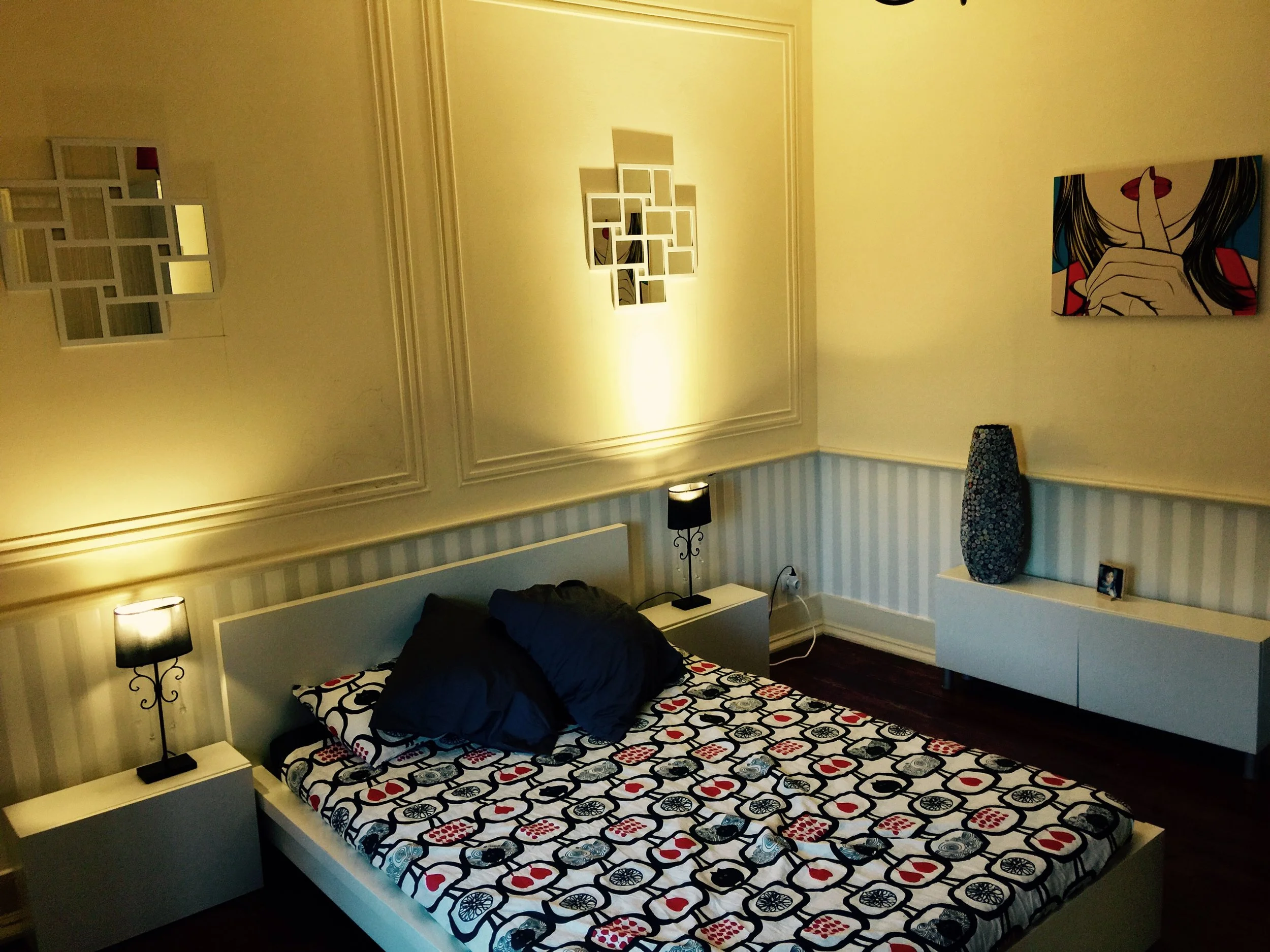 Quarto com cama de casal, duas luminárias de cabeceira, quadro com arte moderna na parede, e uma grande vasilha decorativa azul. O ambiente tem paredes com detalhes de moldura e uma parede com papel de parede listrado azul claro.