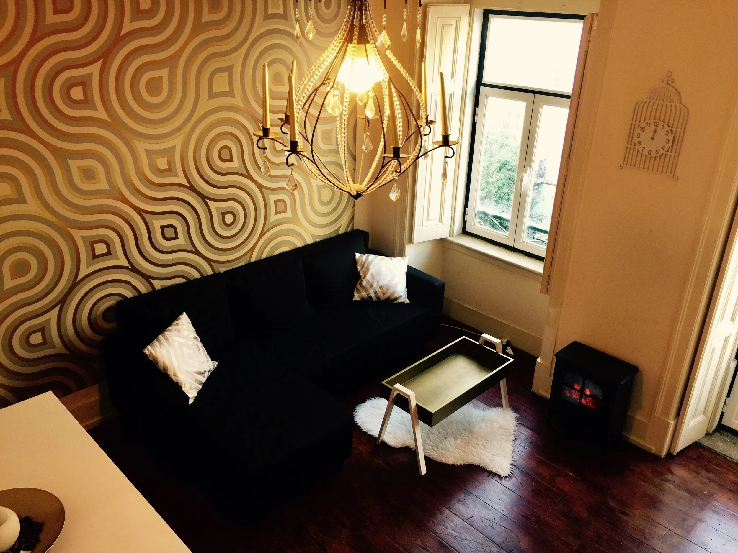 Sala de estar com sofá preto, mesa de centro, tapete branco, janela grande, lustre decorativo com candelabros, parede com papel de parede com desenho colorido, e uma lareira elétrica.