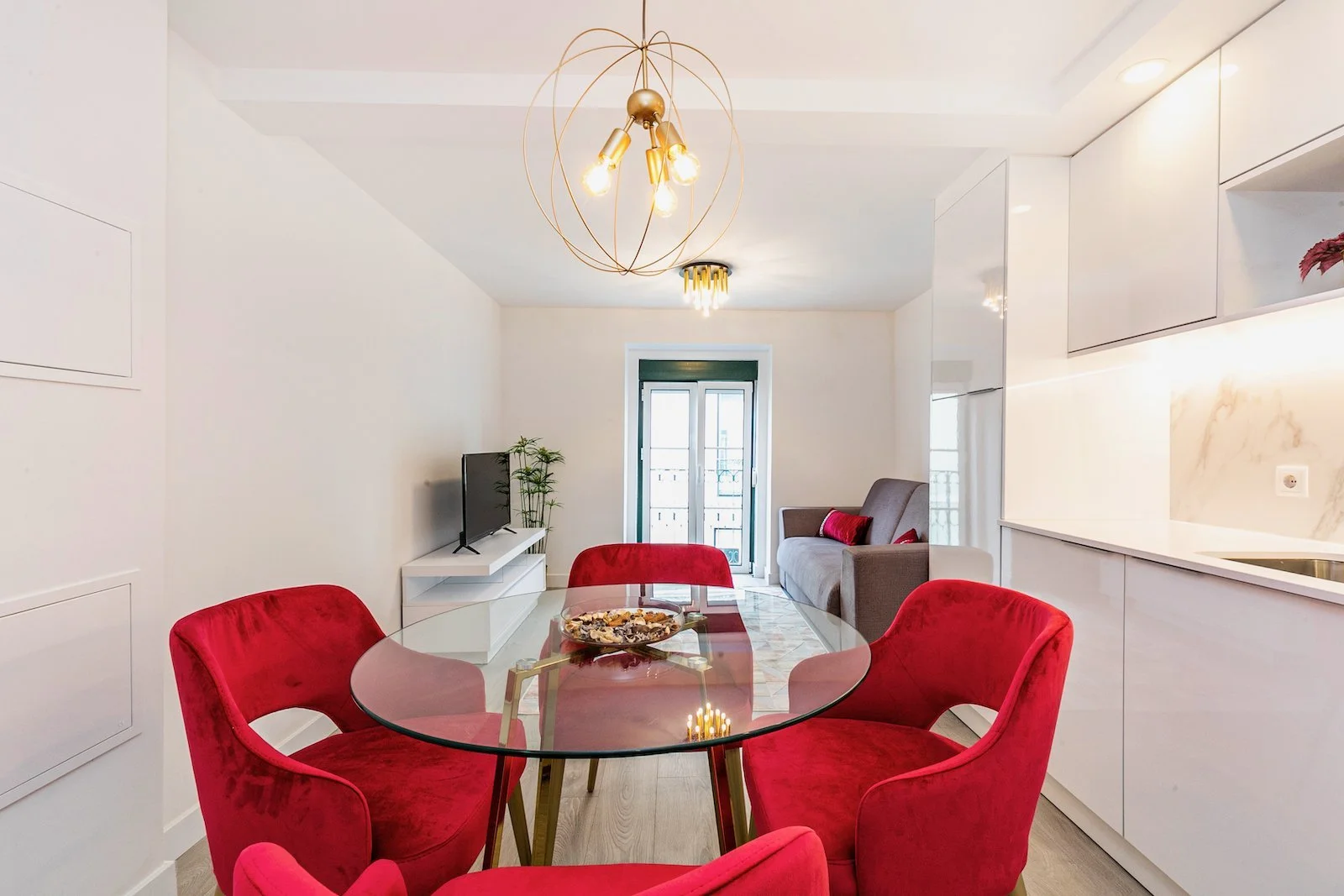 Sala de jantar com mesa de vidro, seis cadeiras vermelhas, sofá cinza, televisão, plantas, iluminação moderna e portas de vidro ao fundo.