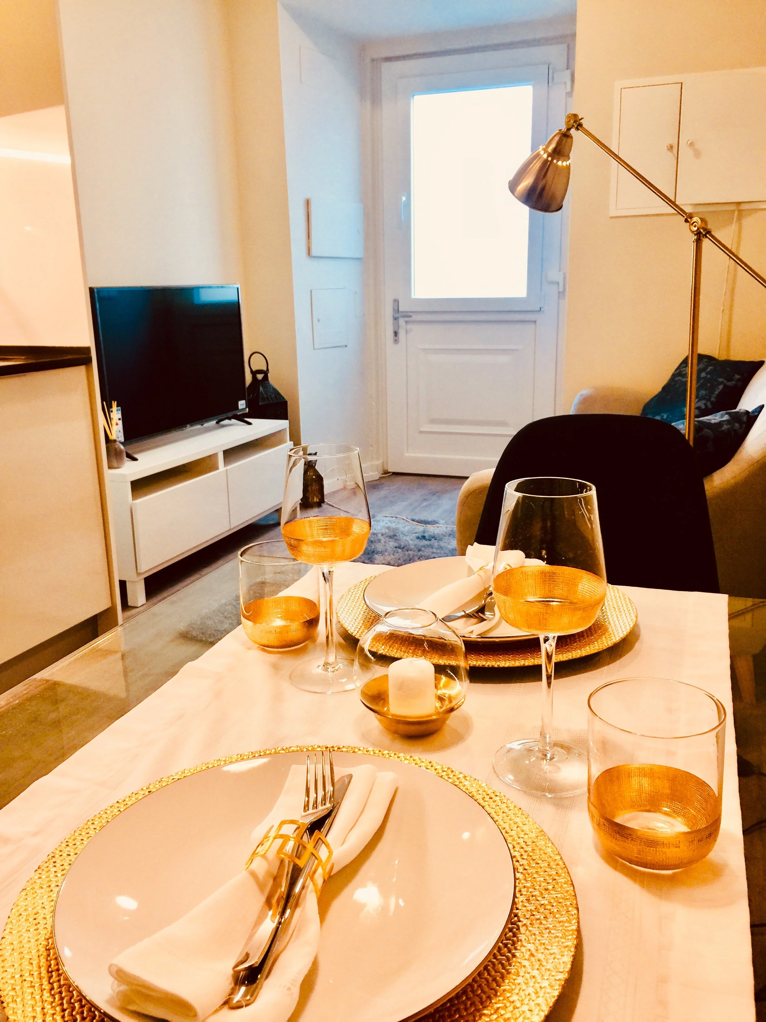 Mesa de jantar decorada com pratos, copos de vinho, taças e velas, em uma sala com sofá, televisão e porta de vidro ao fundo.