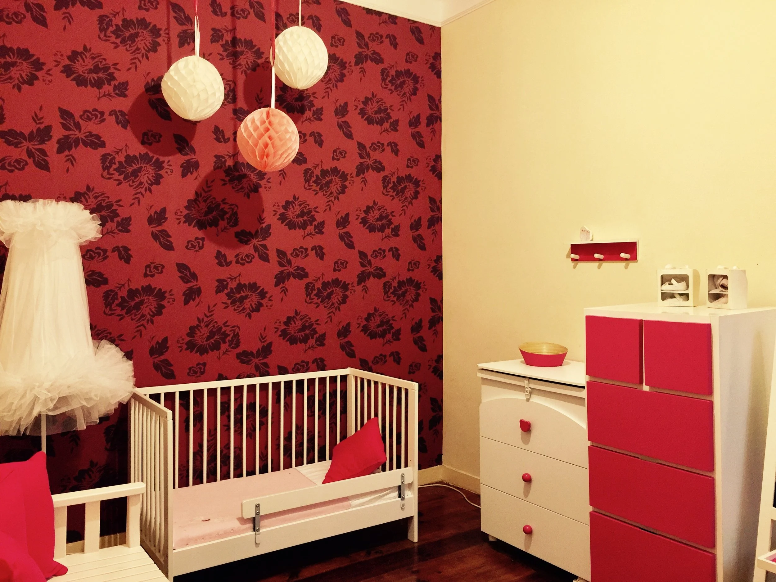 quarto de bebê com parede de destaque vermelha com estampa de flores, berço branco com almofada rosa, cômoda branca com portas vermelhas, decoração rosa e branca, luminárias de papel penduradas no teto.