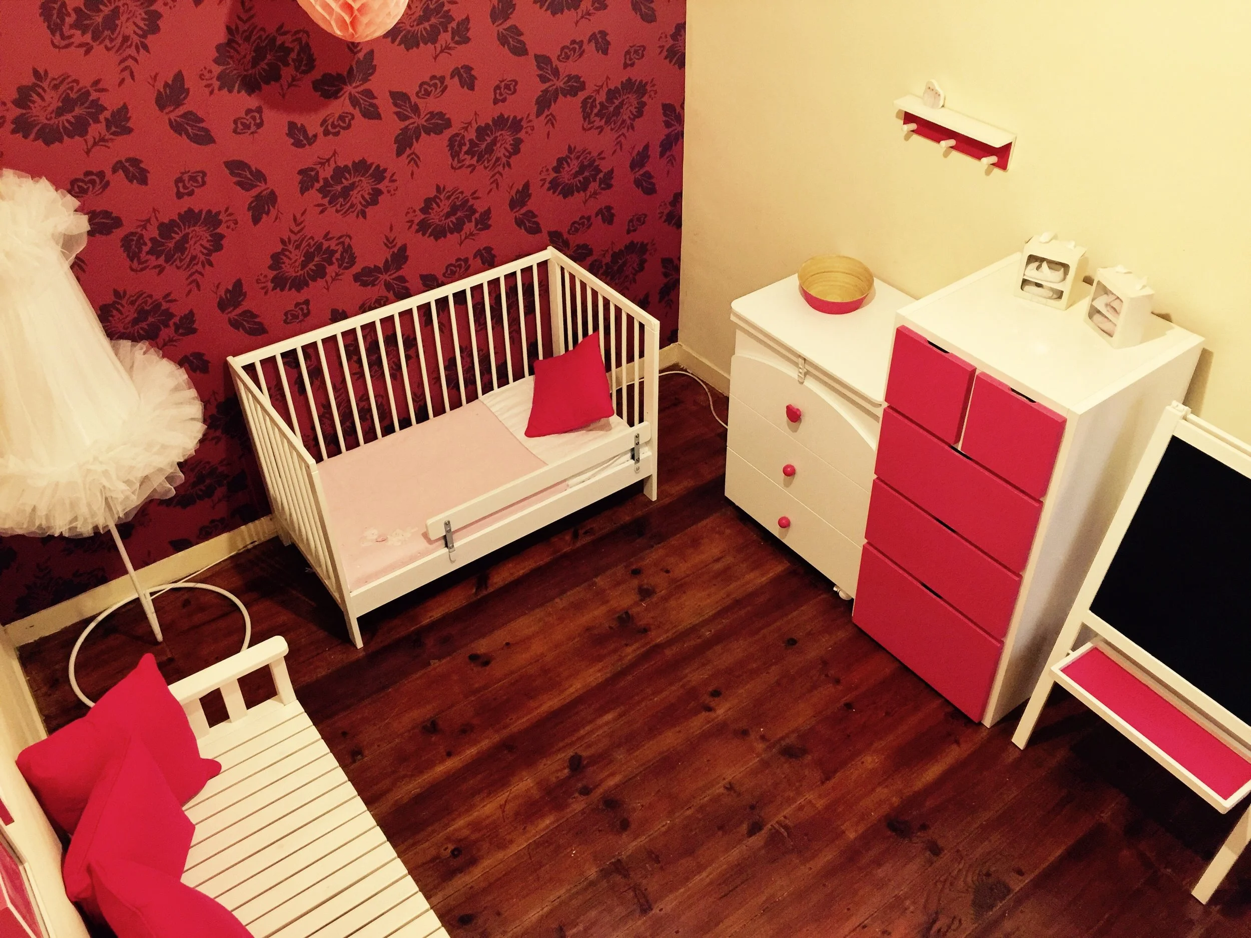 Quarto de bebê com orquídea em rosa, berço branco, poltrona branca com almofadas rosa, cômoda branca com gavetas rosas, prateleira branca com detalhes rosas, cadeira de madeira com assento rosa, parede com papel de parede floral em tons rosa e vermelho.