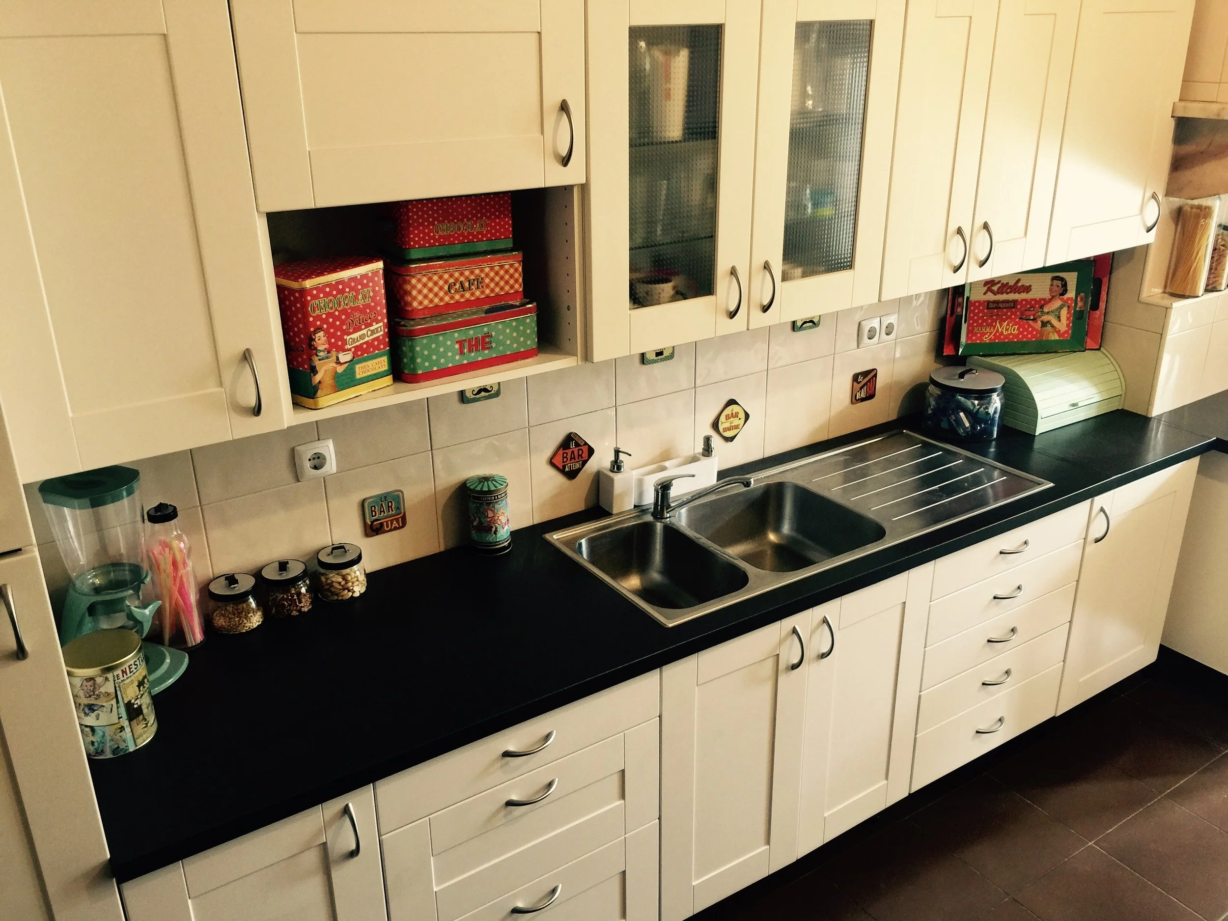 Cozinha com armários brancos, bancada preta, pia dupla de aço inox, utensílios, potes, caixas de alimentos decorativas, eletrodomésticos e objetos de decoração nas paredes.