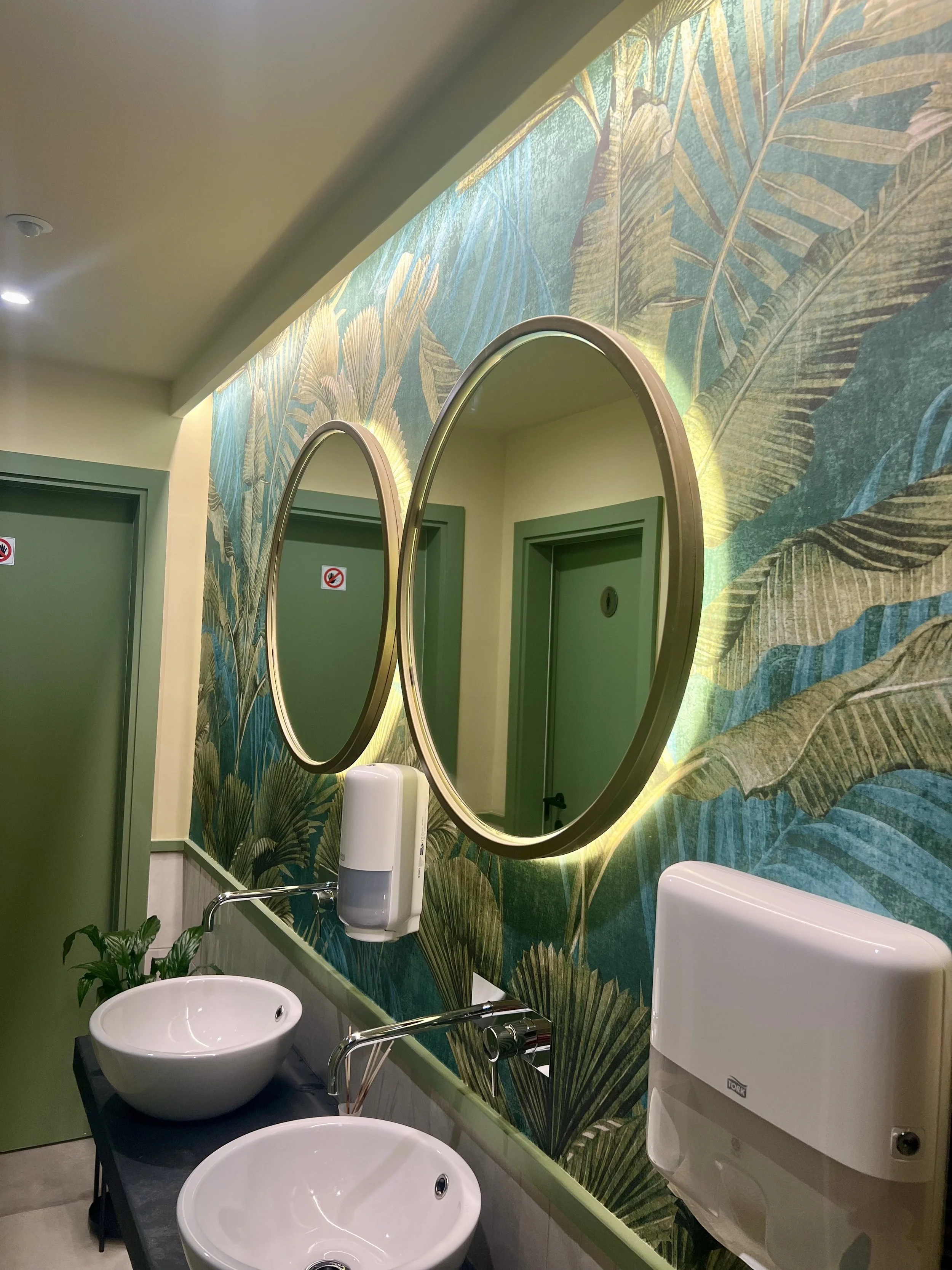 Banheiros com paredes decoradas com papel de parede de folhas tropicais, dois espelhos redondos com iluminação ao redor, duas pias brancas com torneiras modernas, dispenser de sabonete e papel toalha nas paredes