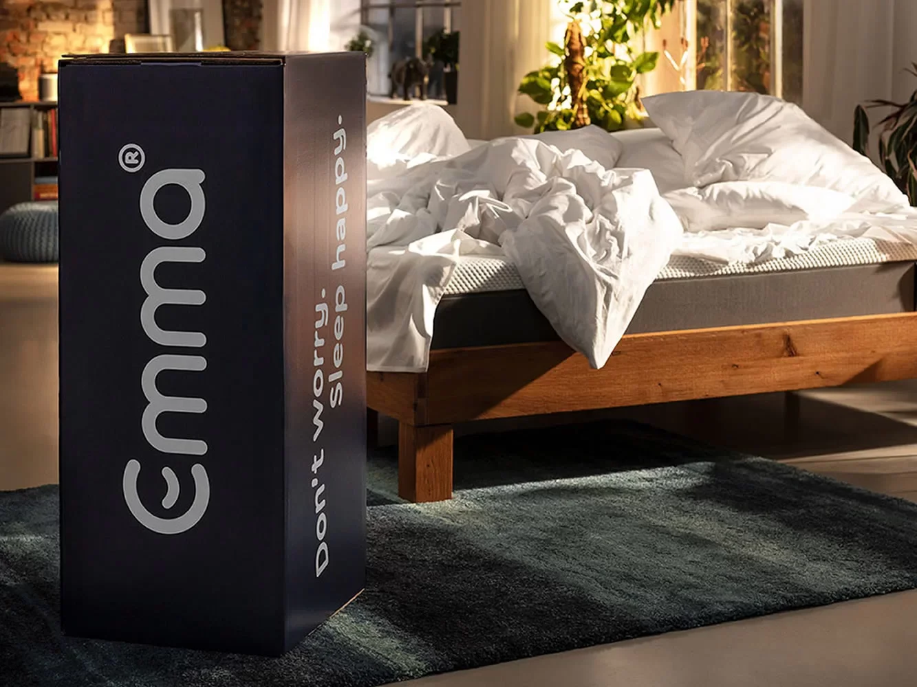 Cama desarrumada com roupa de cama branca, ao lado de uma caixa de travesseiros Dyson. Quarto com decoração de plantas e móveis de madeira.