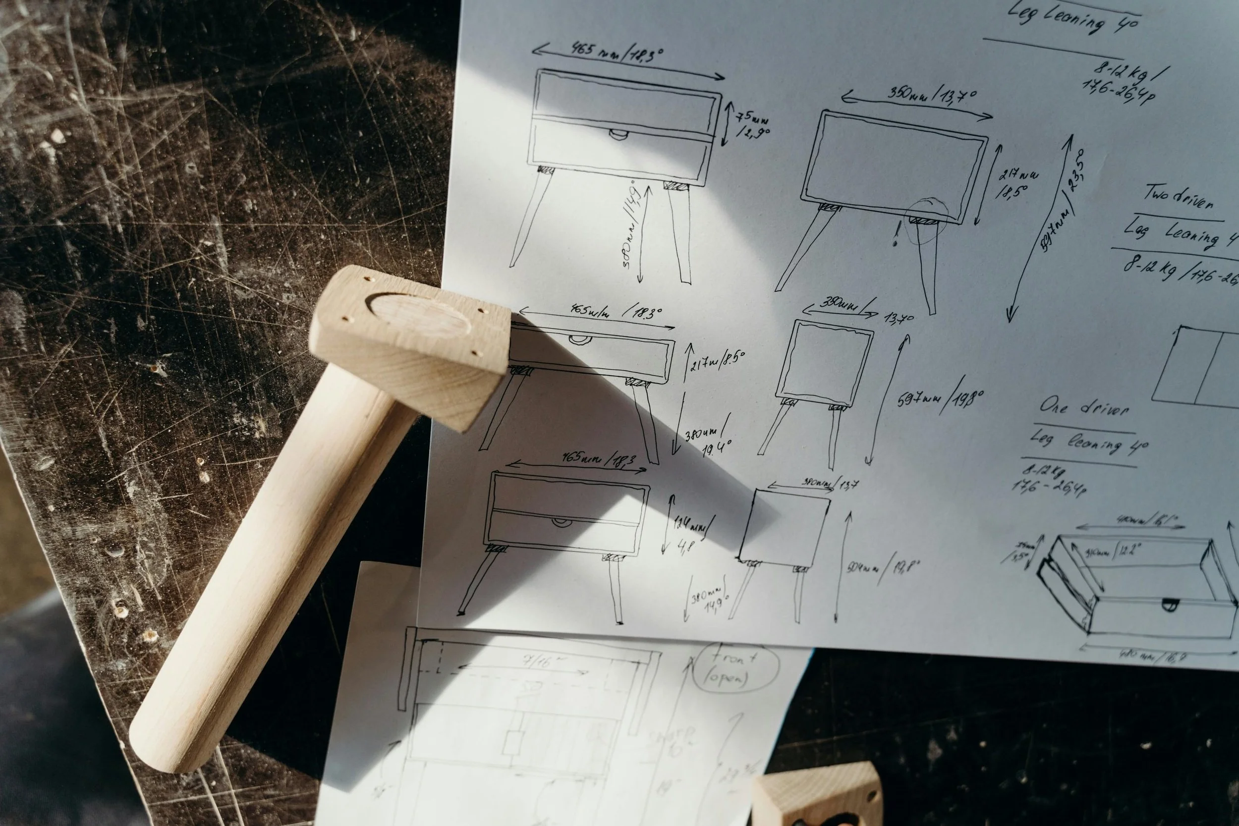 Desenhos técnicos de uma bancada com gaveta, com medidas e detalhes, colocados sobre uma mesa de madeira escura. Há também um pedaço de madeira com furo na imagem.