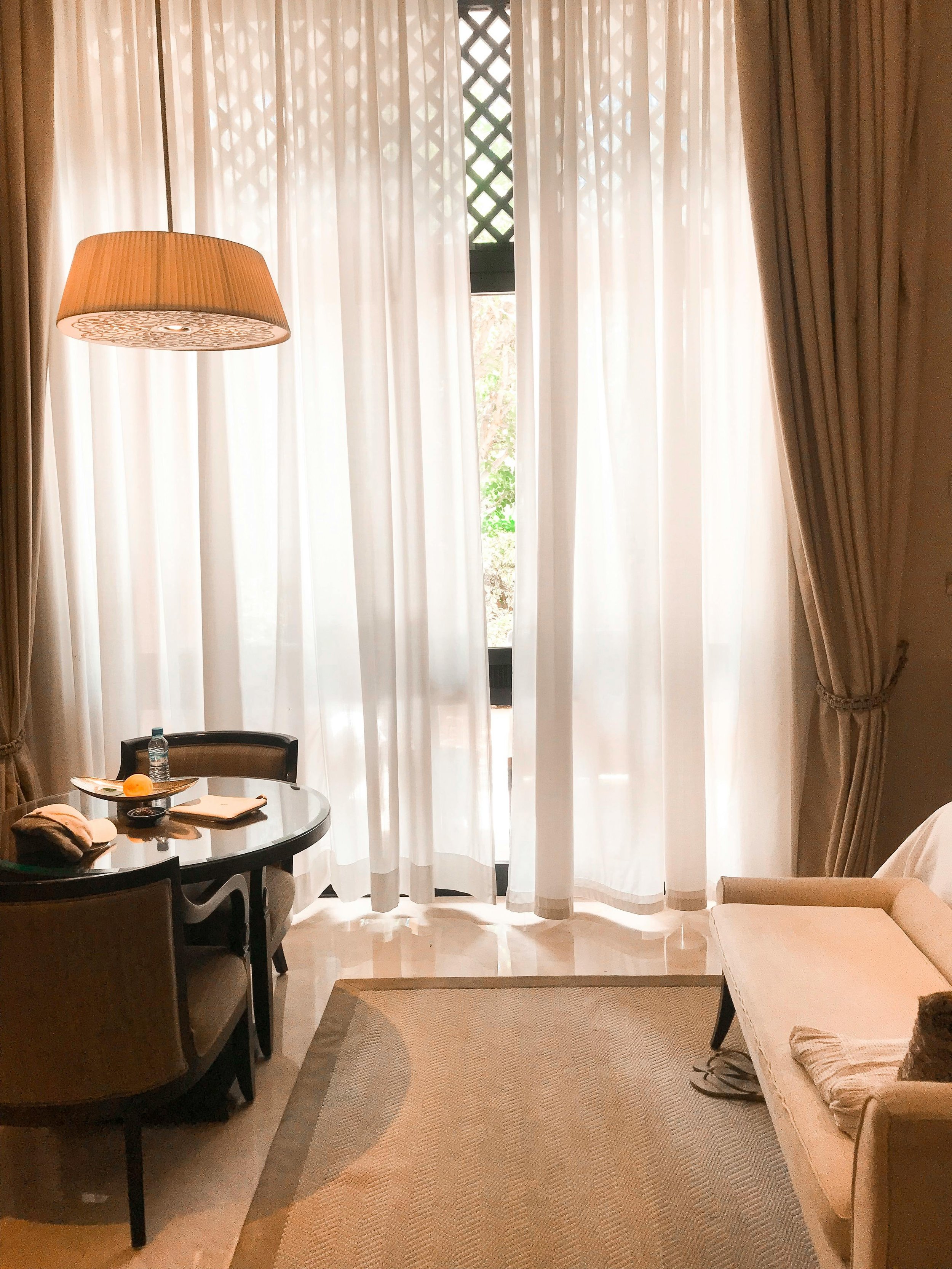 Janela com cortinas brancas e persianas, com uma sala de estar ao fundo, contendo uma mesa com cadeira, uma garrafa de água, um e-book, uma toalha, uma cômoda e um espelho.